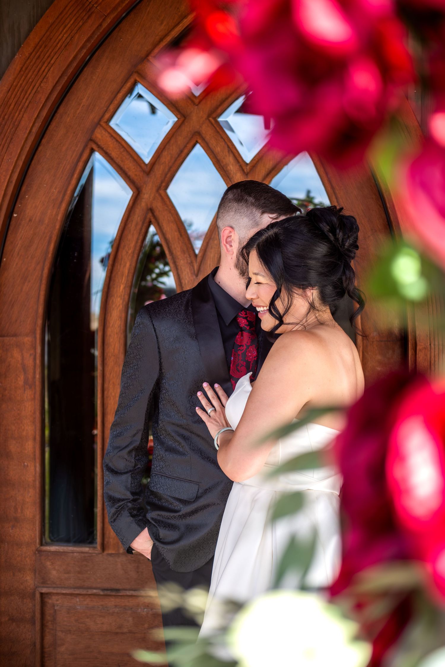 Bride and groom share intimate moment framed by ornate wooden arched church doors and red flowers.
