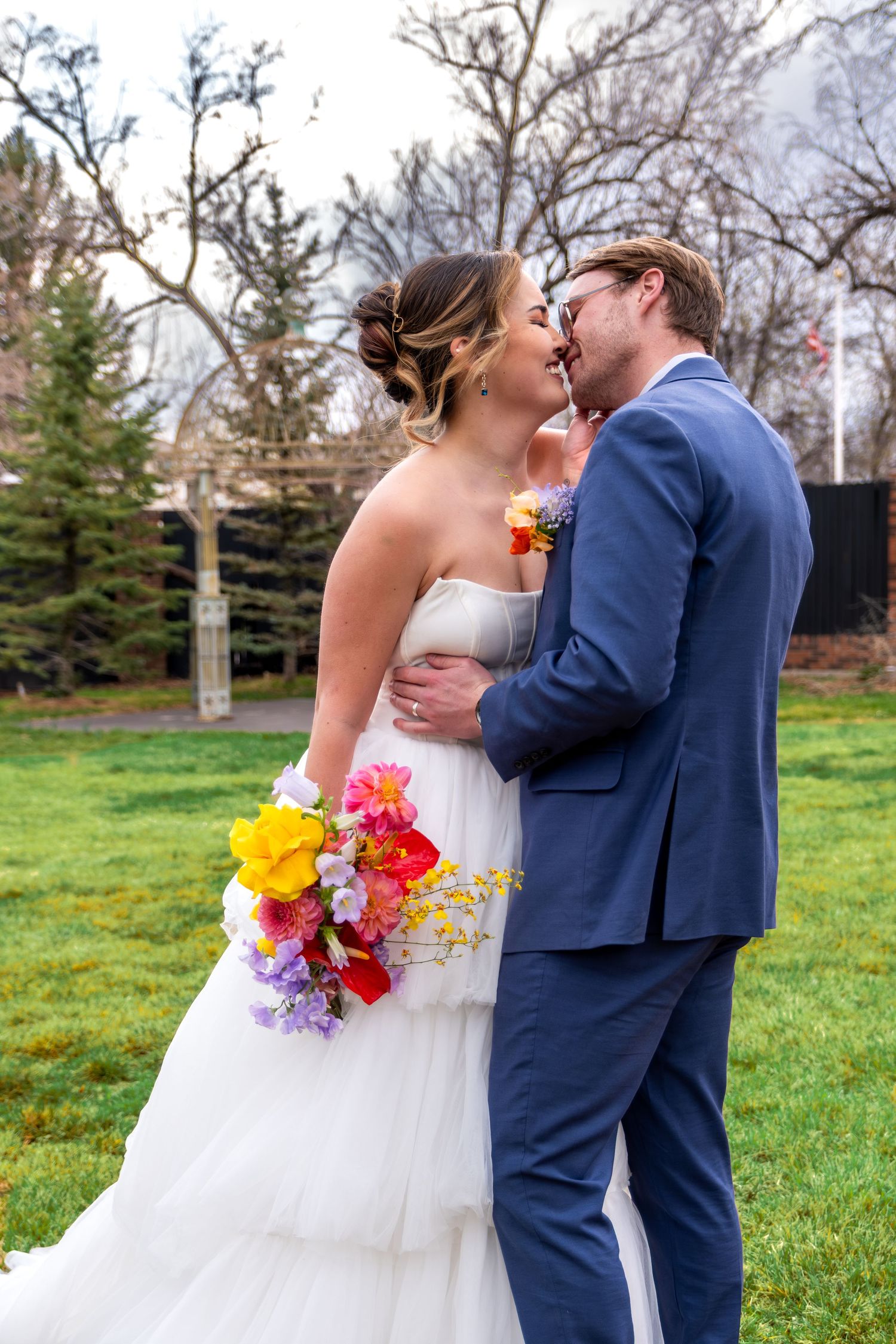 Bride and groom sharing romantic almost-kiss, holding vibrant multicolored bouquet in outdoor garden setting.