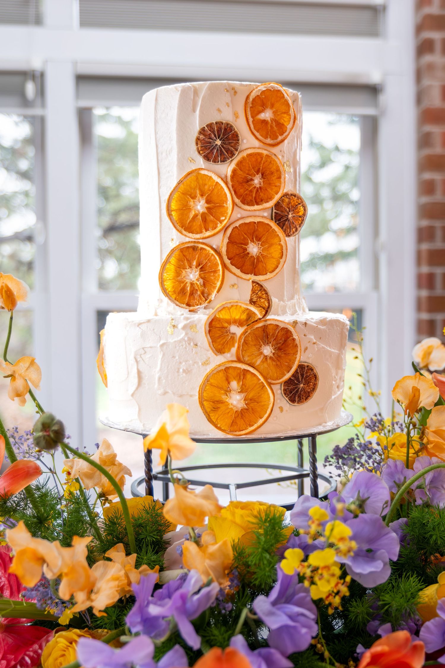 Elegant two-tier white wedding cake decorated with dried orange slices surrounded by colorful flowers.