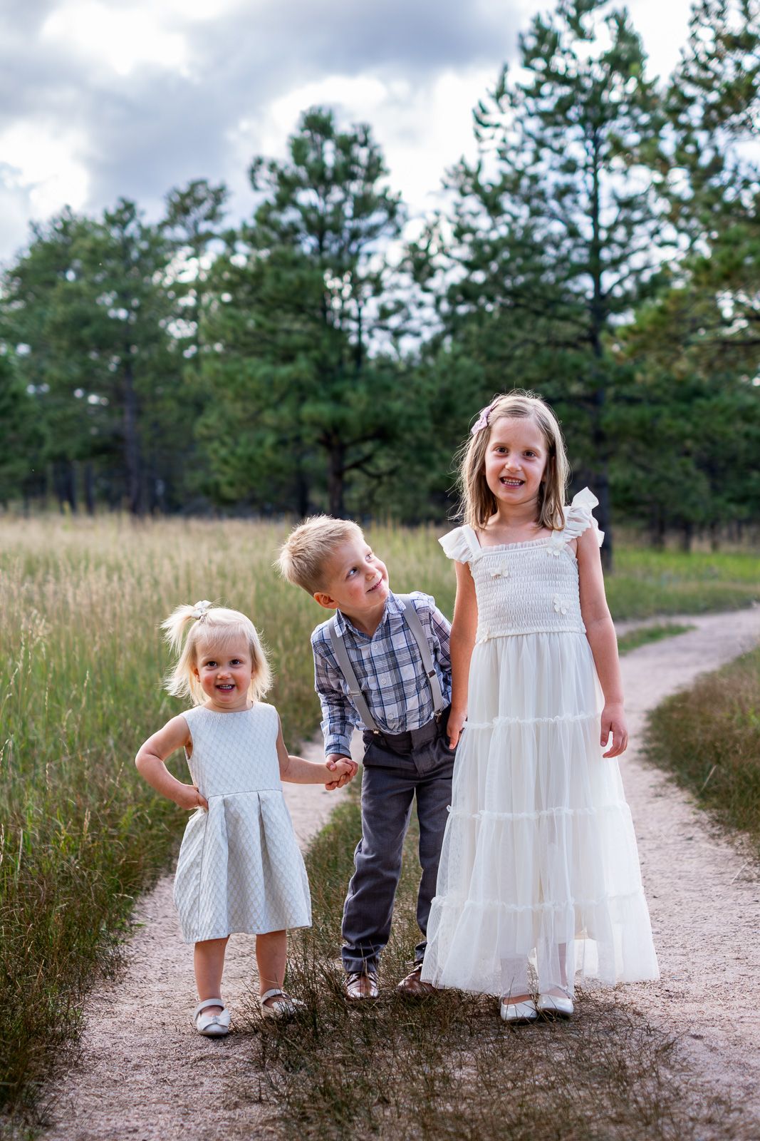 Three siblings in coordinated outfits smile together on a dirt path surrounded by tall grass.