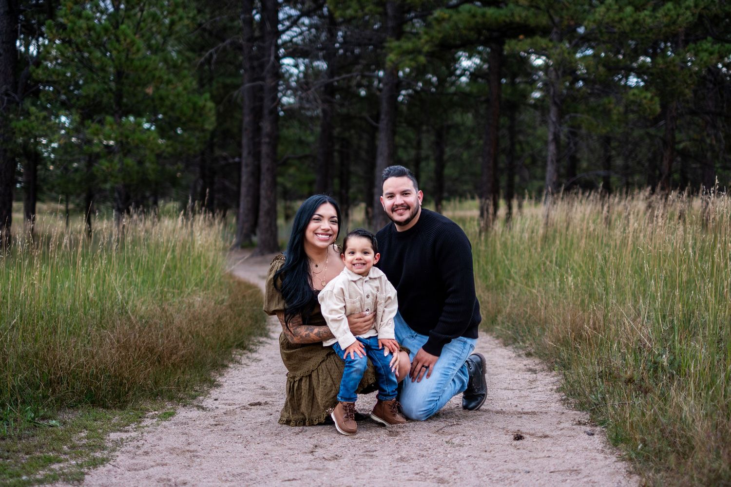 Family of three posing together on a sandy path through a pine forest on a cloudy day.