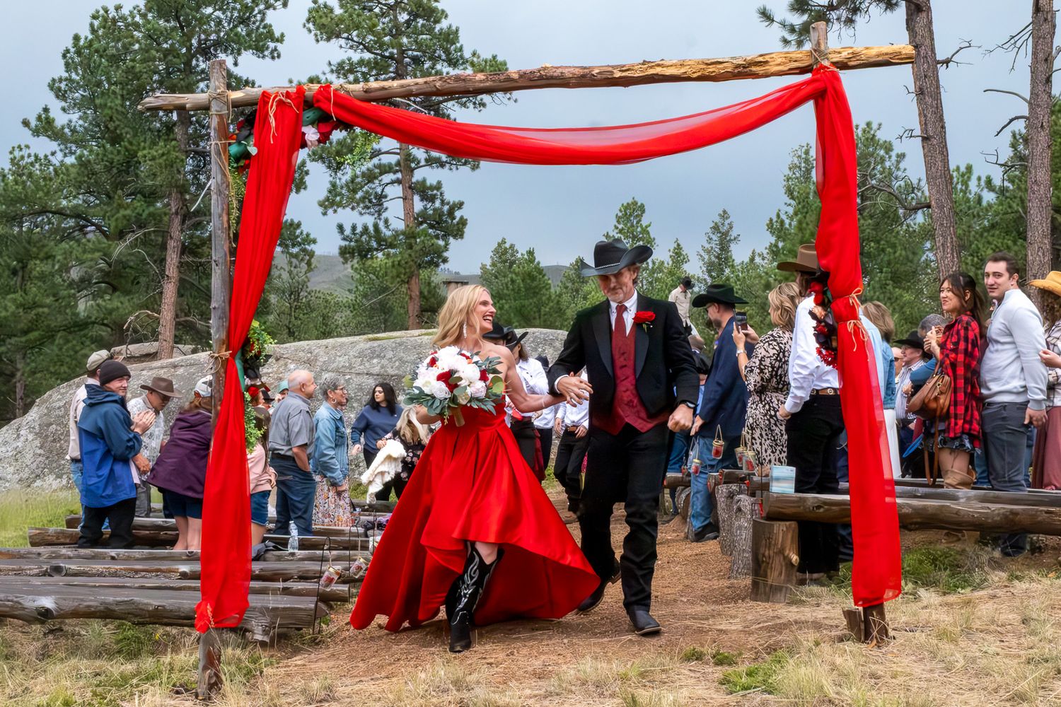 Western-themed wedding couple exits ceremony under red-draped rustic log arch surrounded by guests outdoors.