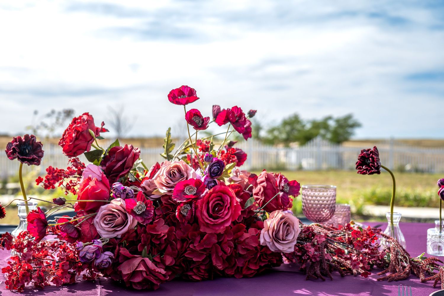 Lush red and pink floral centerpiece with roses and ranunculus on purple table under open sky.