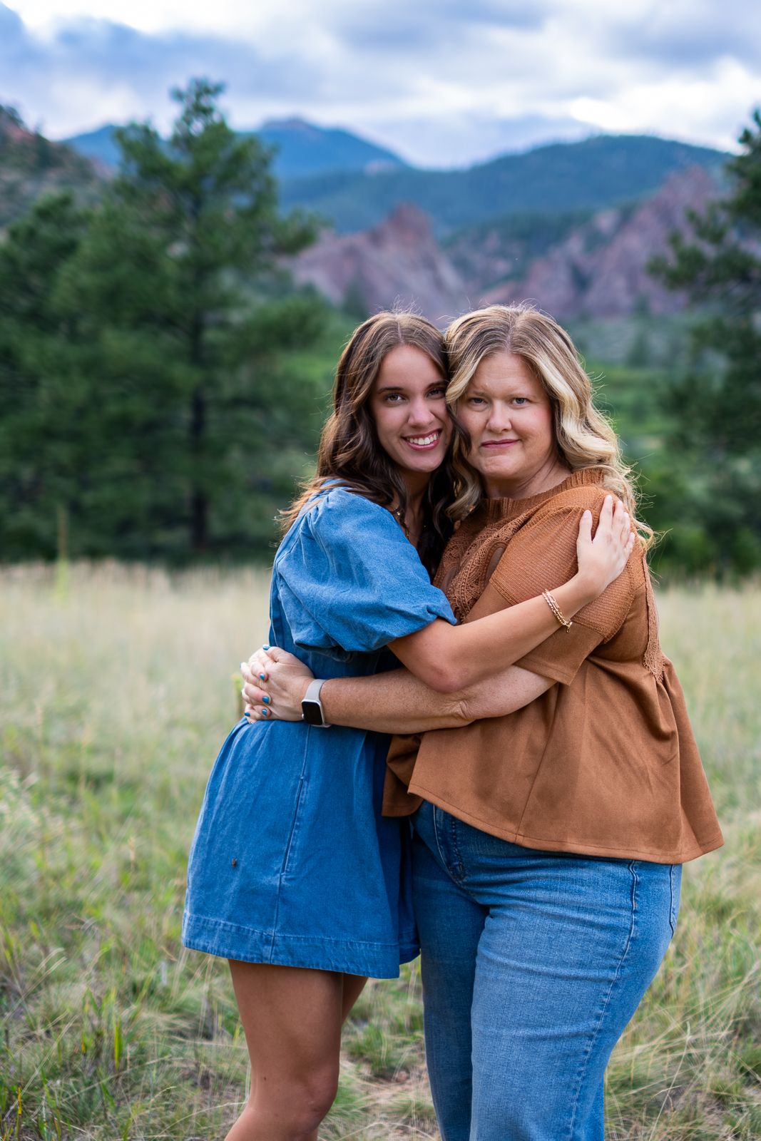 Two women hugging and smiling in a mountain meadow with pine trees and rocky peaks in Colorado.
