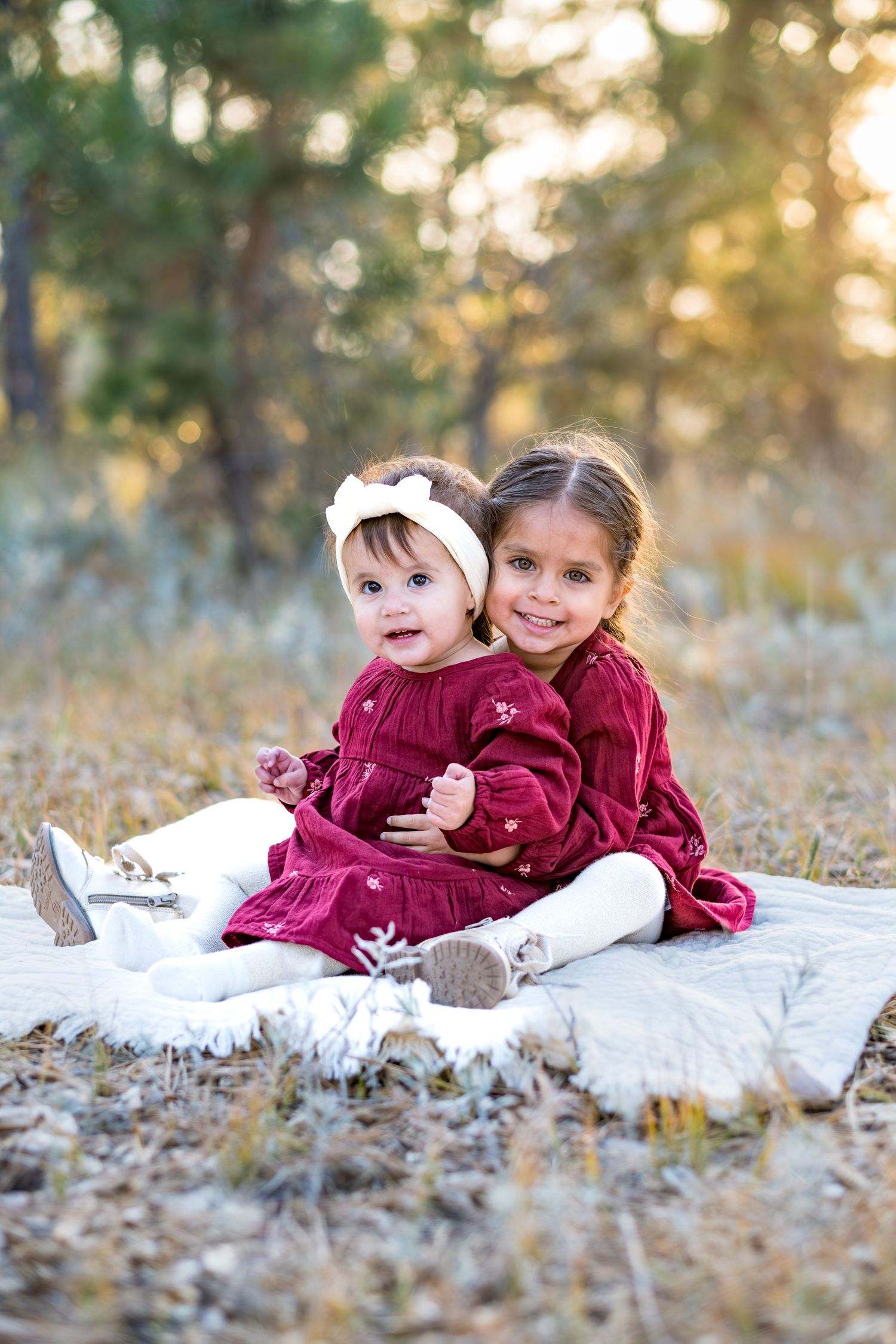 Two young sisters in matching burgundy dresses sitting together on white blanket in autumn forest setting.