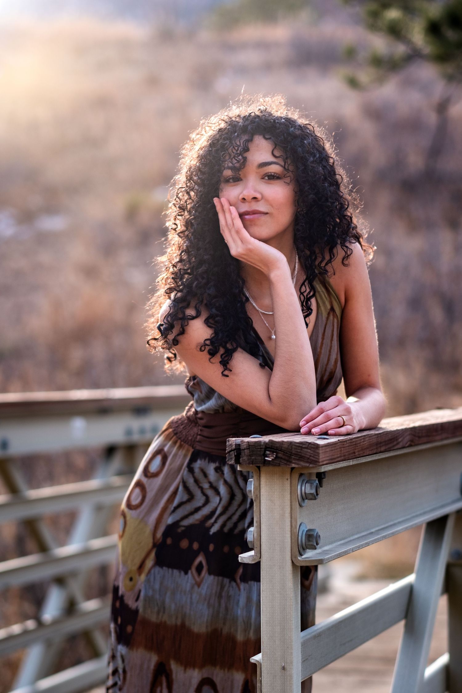 Woman with curly hair in patterned dress leaning on wooden railing in golden sunlight.