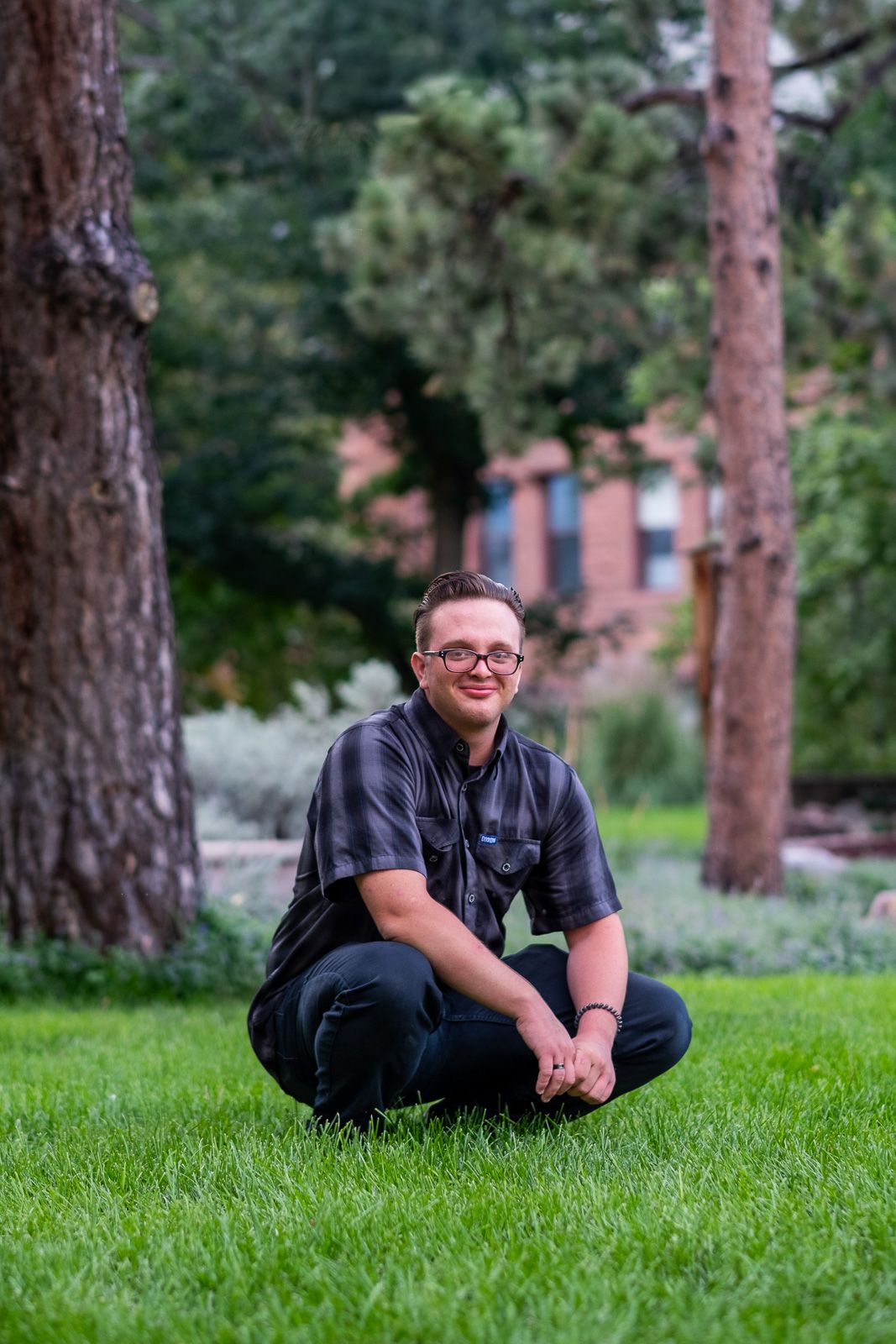 Young man with glasses sitting cross-legged on green grass between tall trees in a park setting.