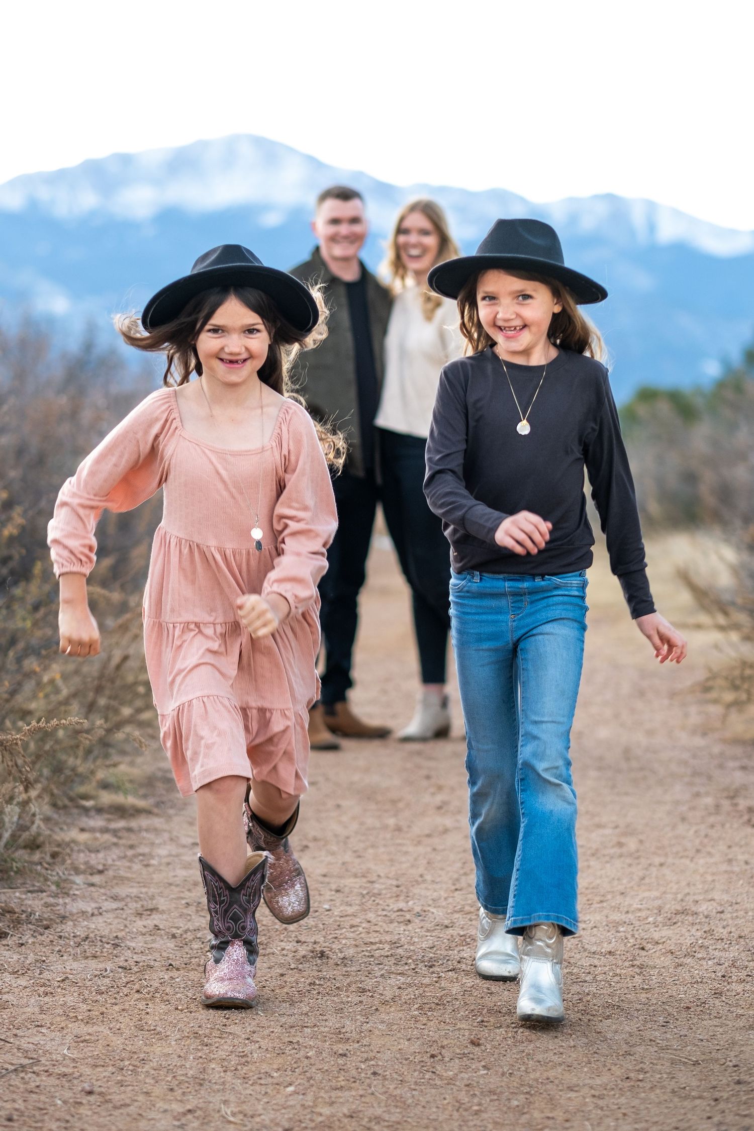 Two young girls in black cowboy hats run happily down a dirt path with their parents and mountains behind them.