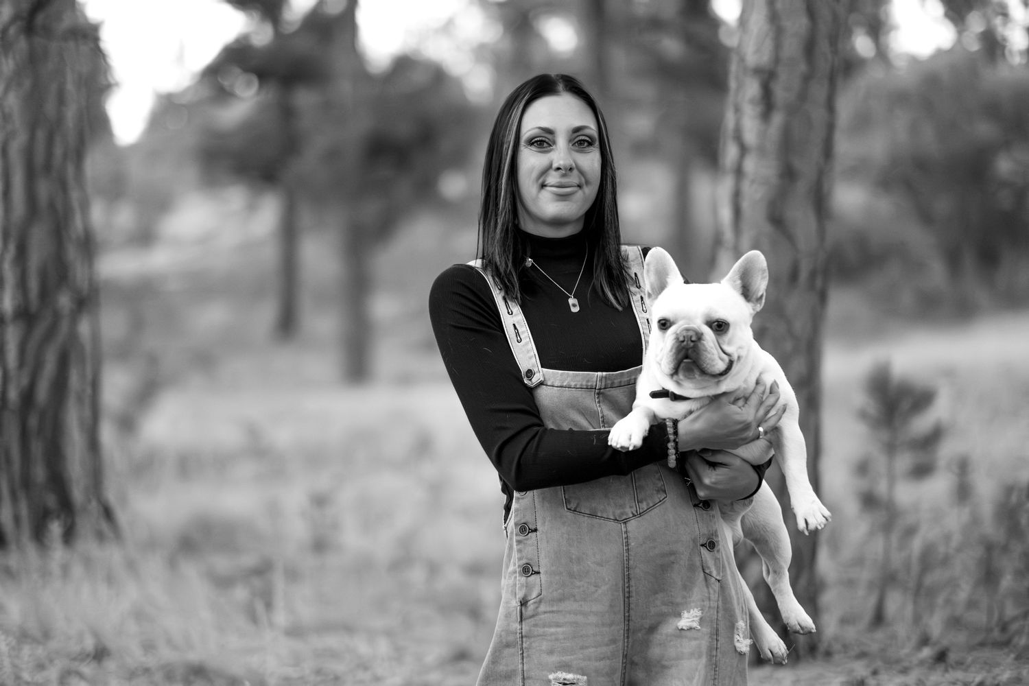 Black and white photo of woman in overalls holding a French Bulldog in a forest setting.