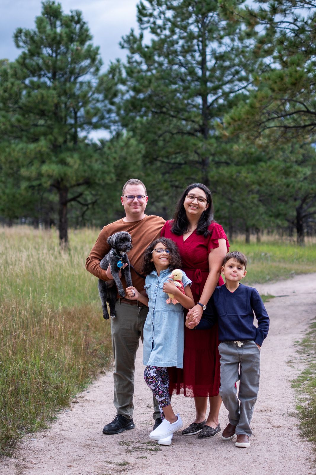 Smiling family with two children and a black puppy posing on a dirt path surrounded by pine forest.