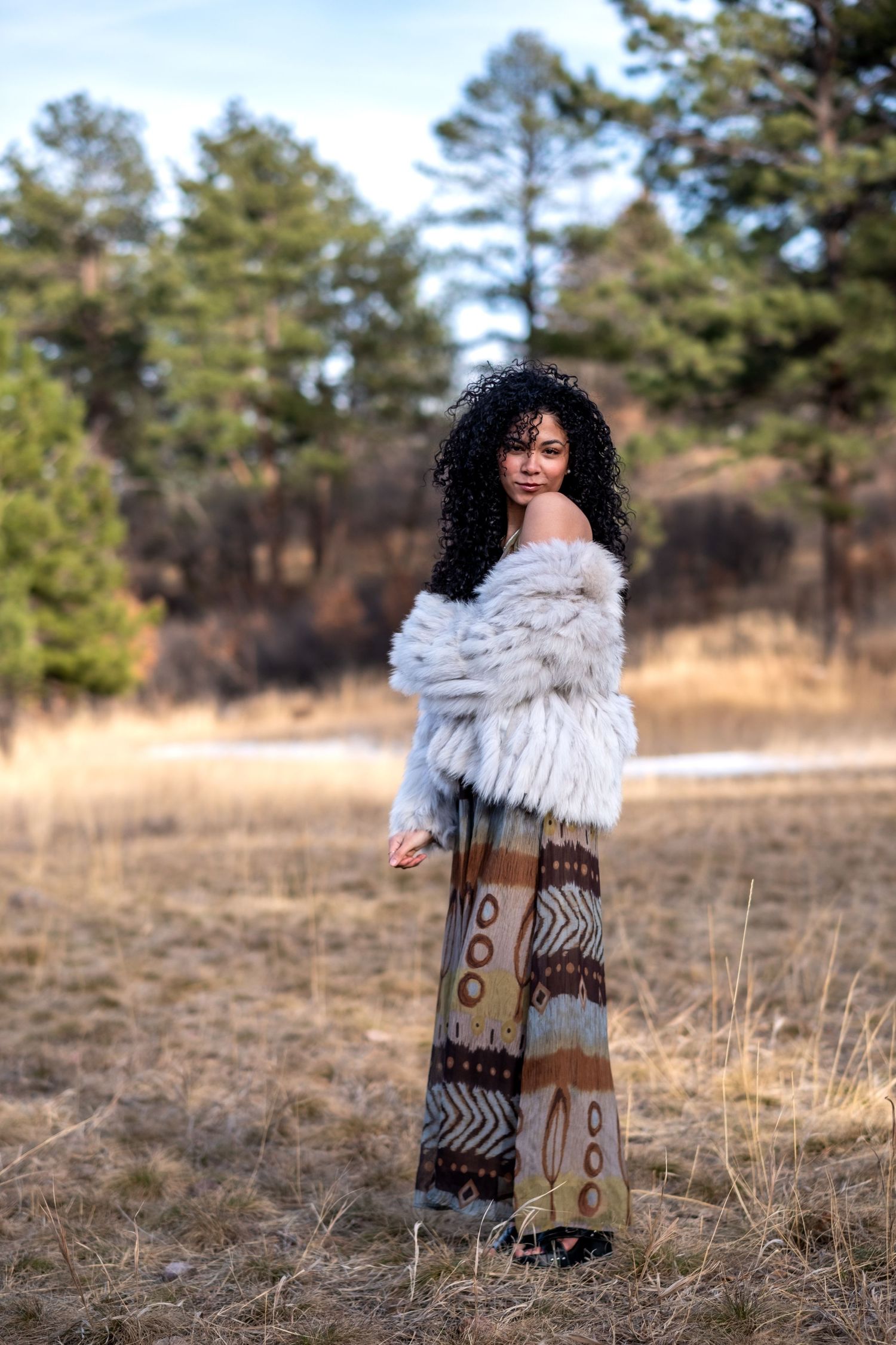 Woman in fur jacket and bohemian wide-leg pants posing in autumn field with pine trees.