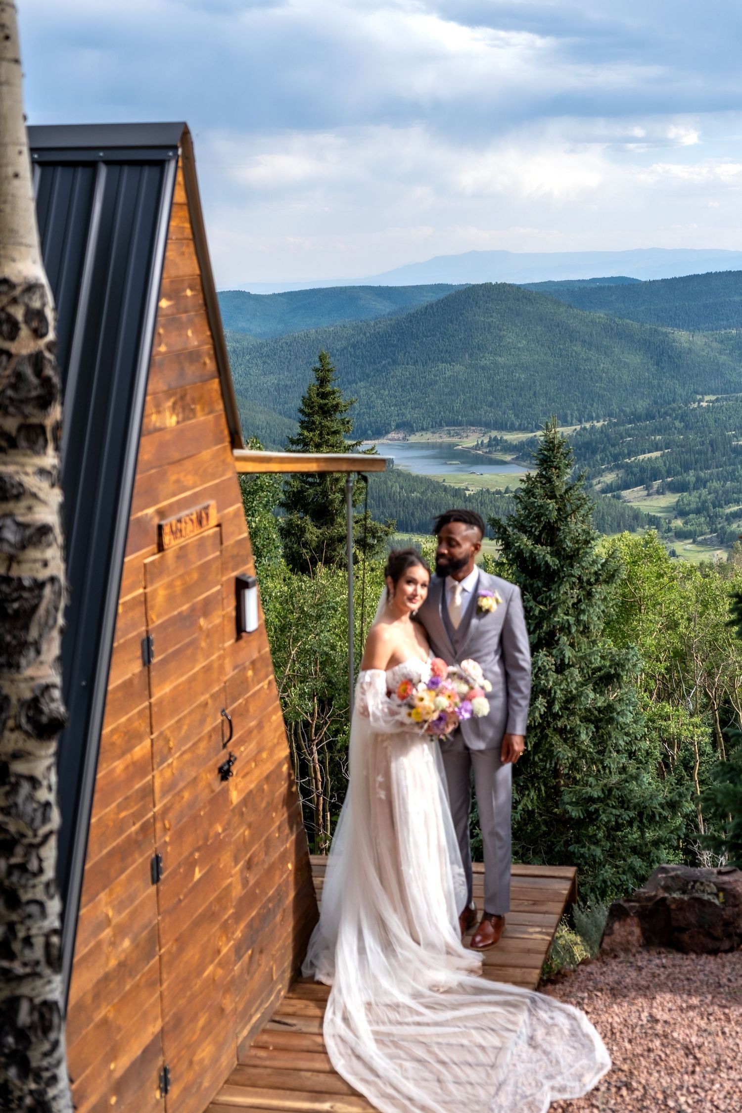 Bride and groom pose together beside wooden A-frame cabin with stunning mountain valley backdrop.