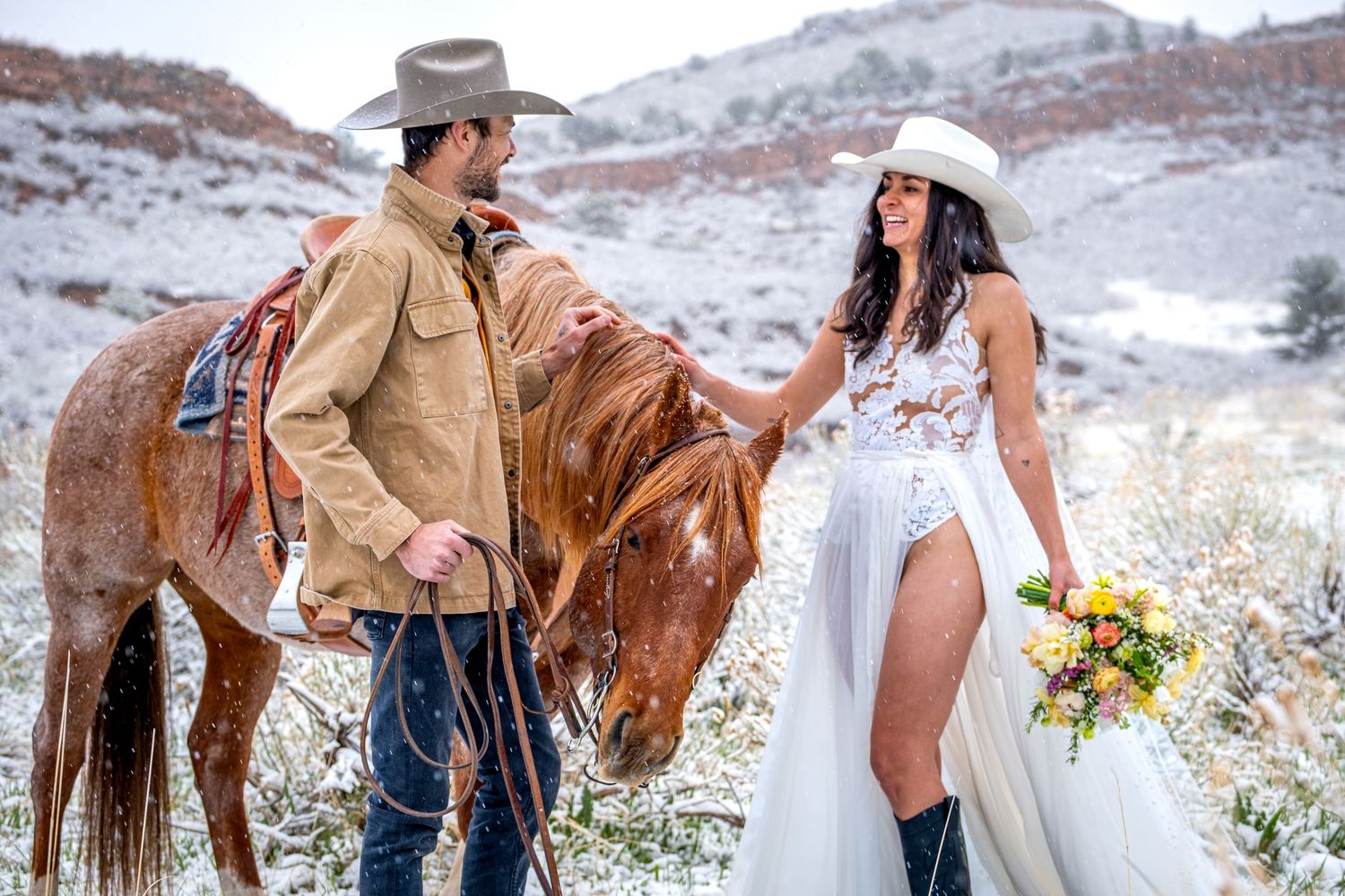 Western wedding couple with chestnut horse in snowy red rock landscape, bride holding colorful bouquet.