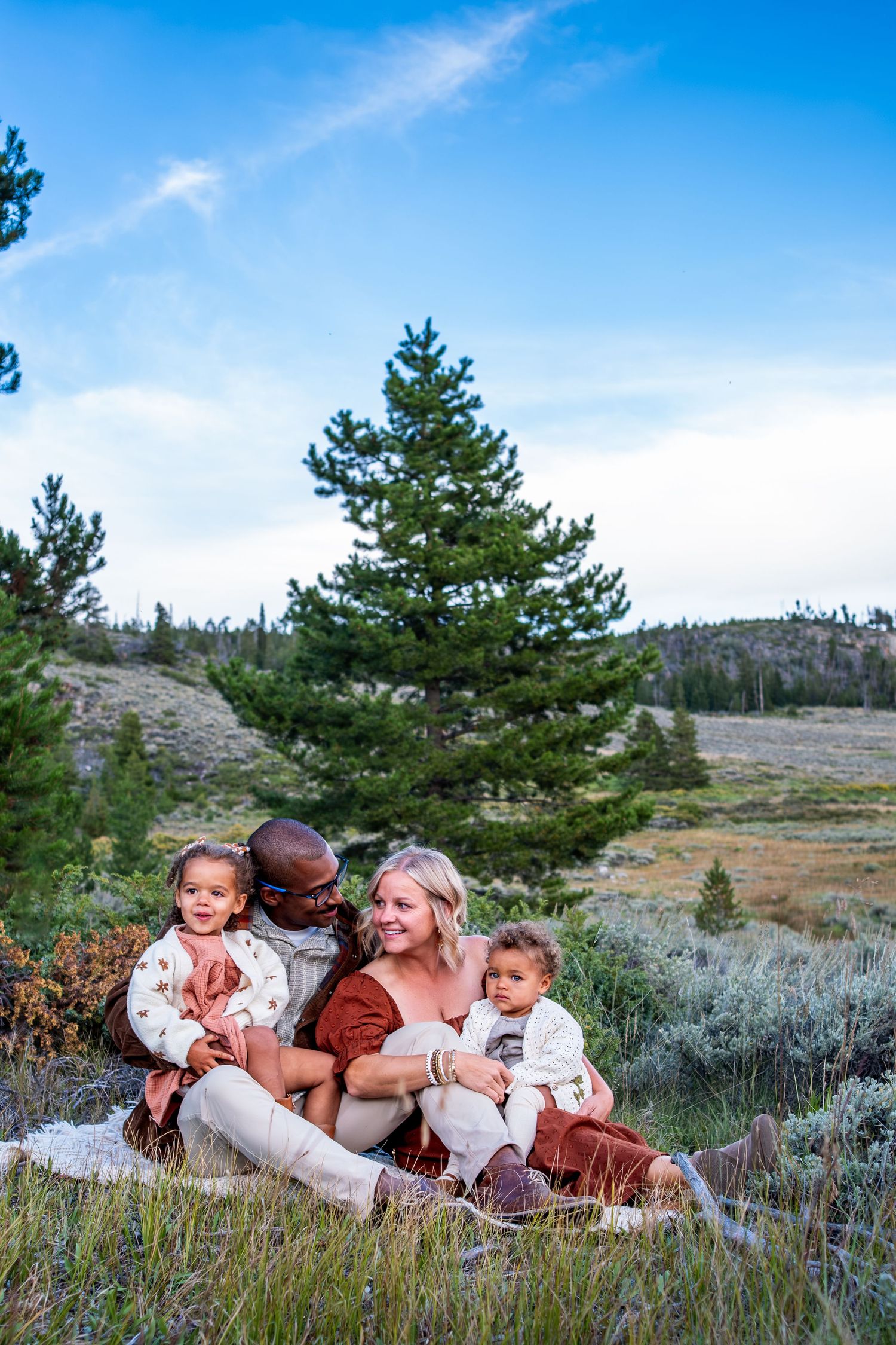 Happy interracial family with two toddlers sitting together in a mountain meadow with pine trees and blue sky.