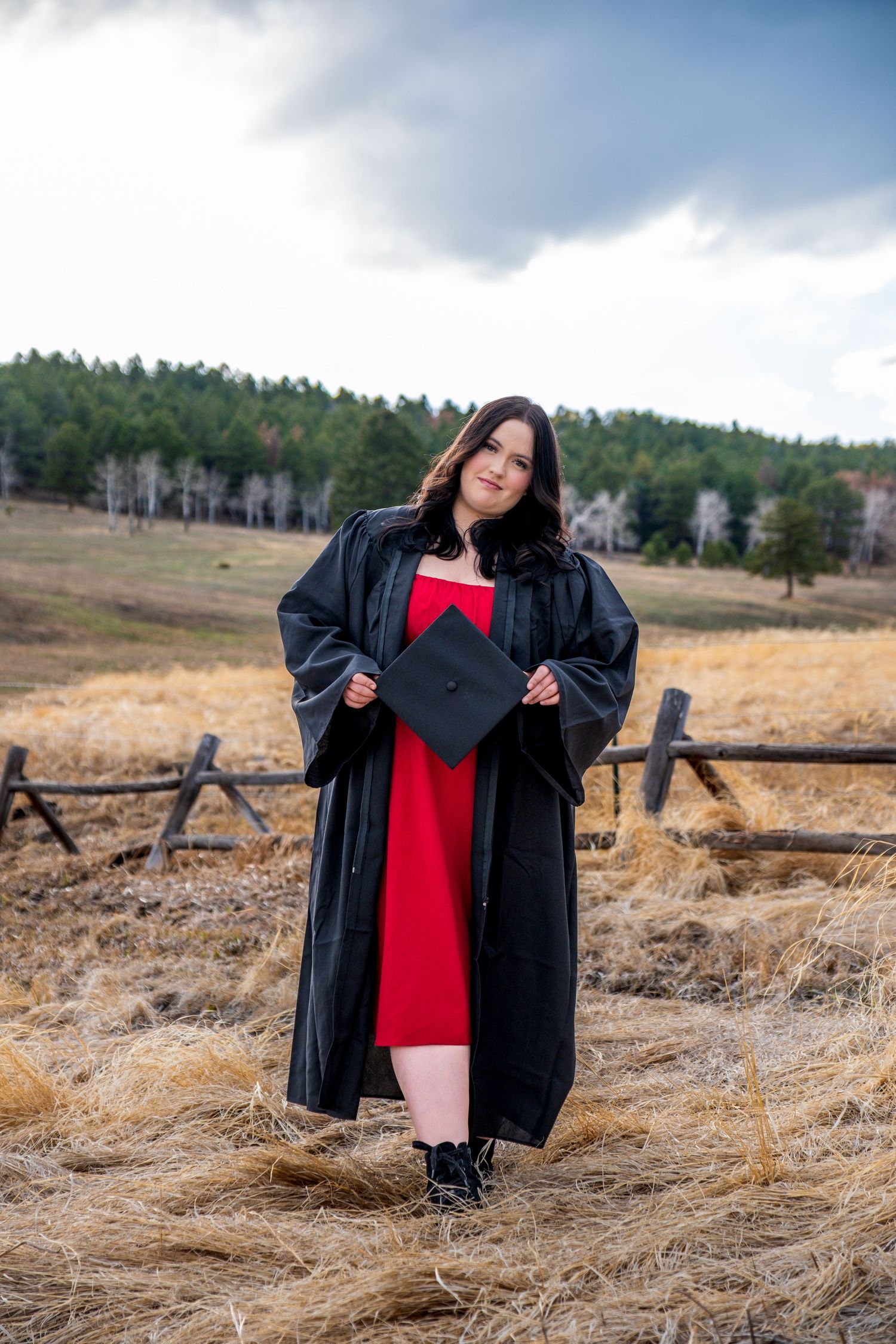 Young woman in black graduation gown holding cap in red dress outdoors in rustic field with wooden fence.