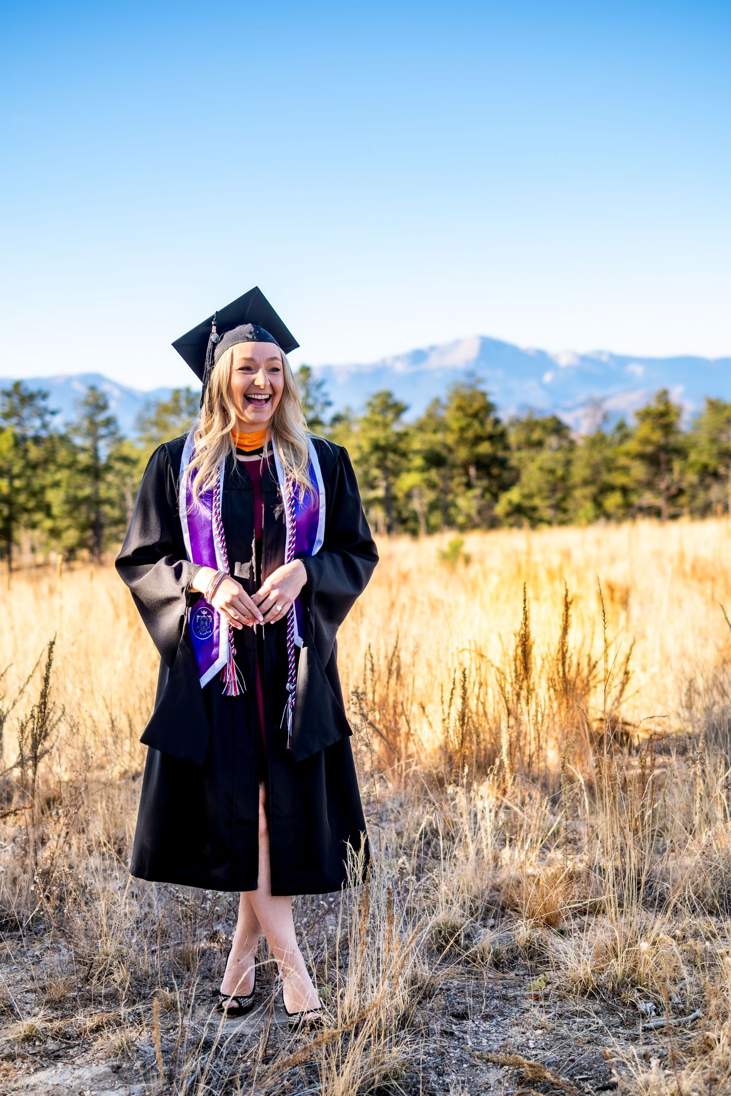 Smiling blonde woman in graduation cap and gown standing in a field with mountains in the background.