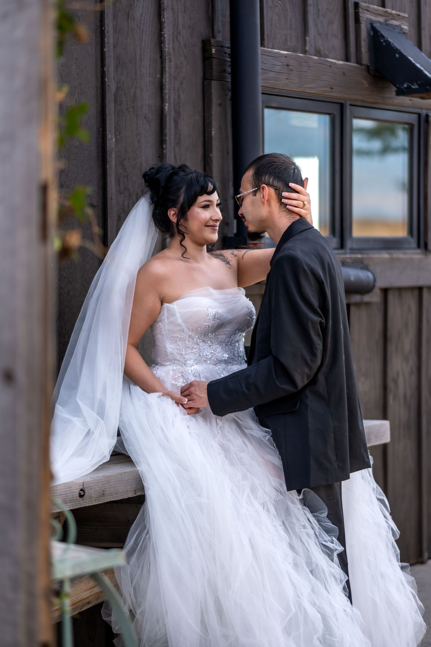 Bride in white strapless tulle gown and groom sharing an intimate moment by a rustic wooden barn.