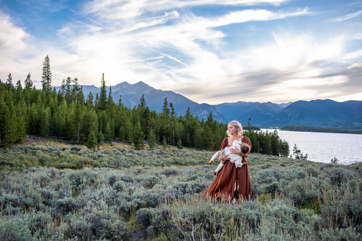 Woman in rust dress holding baby in wildflower meadow with mountain lake and pine trees at sunset.
