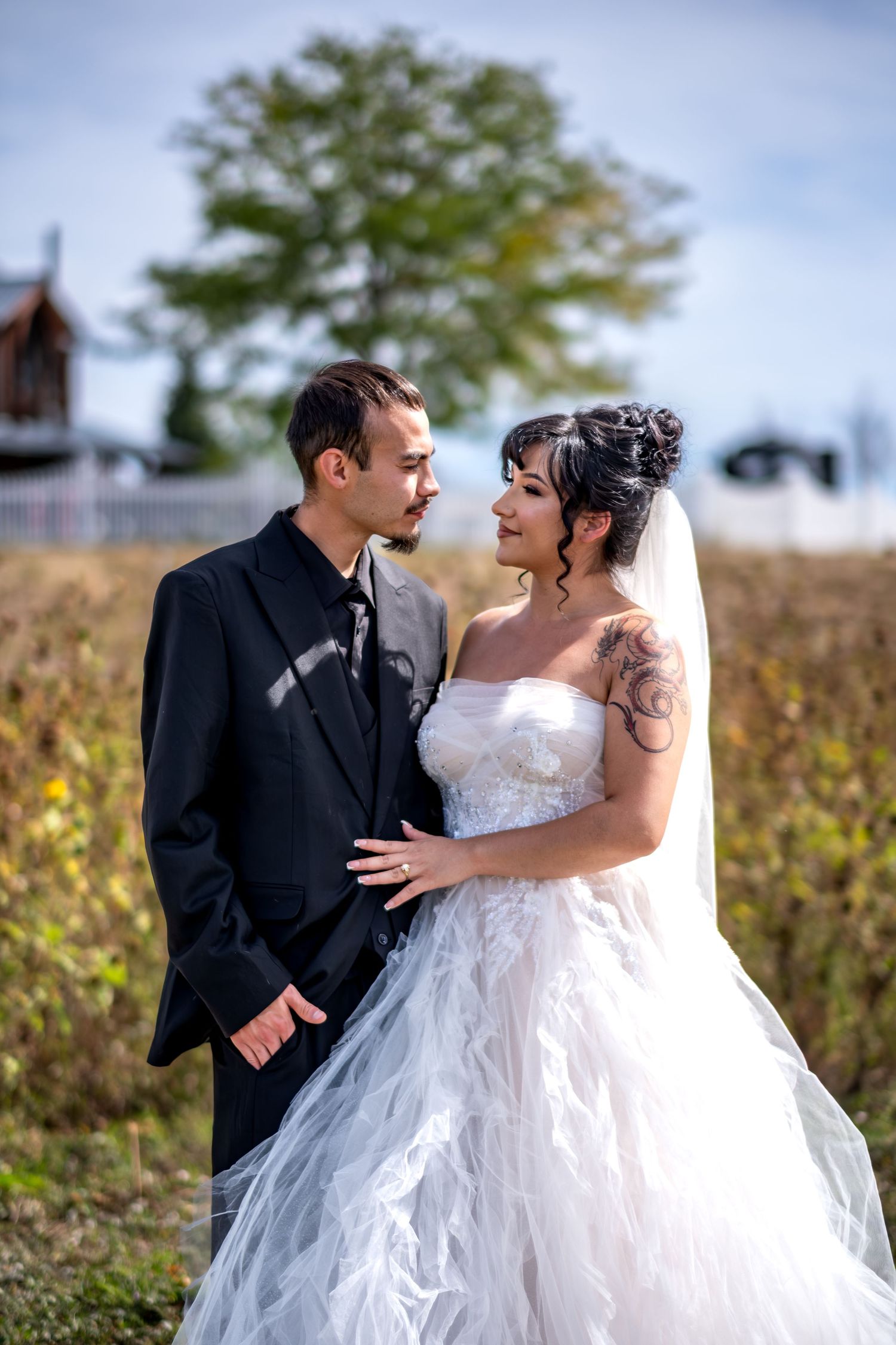 Bride and groom in black suit gazing at each other in an autumn field with trees behind them.