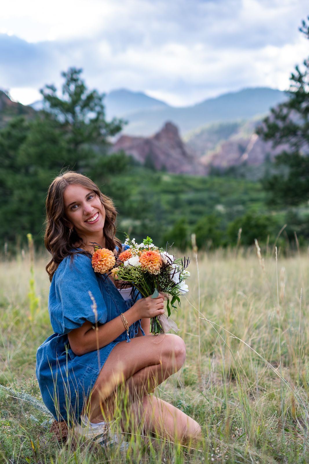 Smiling woman in blue dress kneeling in field holding orange dahlia bouquet near mountains.