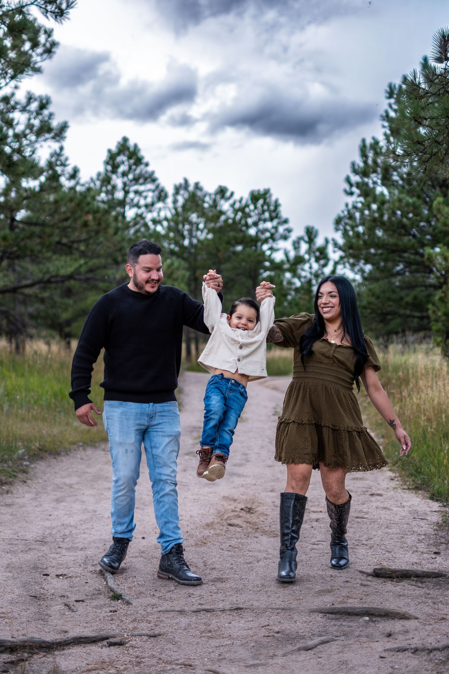 Happy family swinging young child by hands on a dirt path surrounded by pine trees.