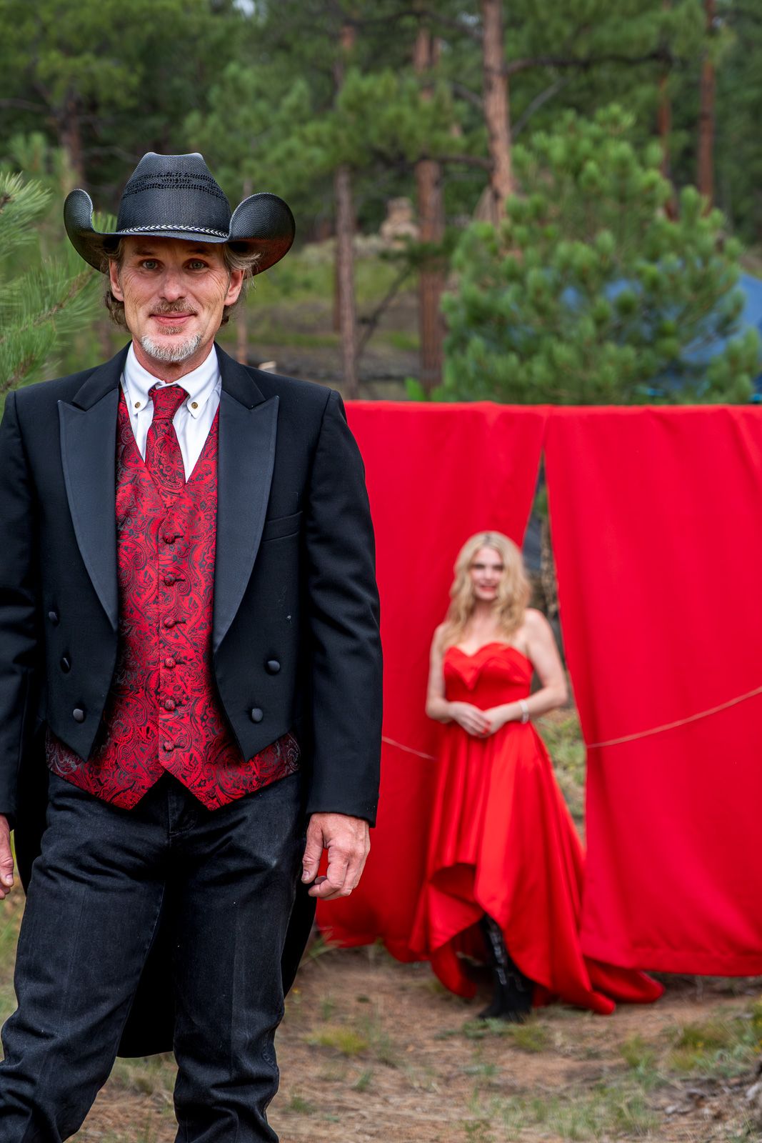 Groom in black tuxedo and cowboy hat stands before red curtain as bride in red gown poses behind him.