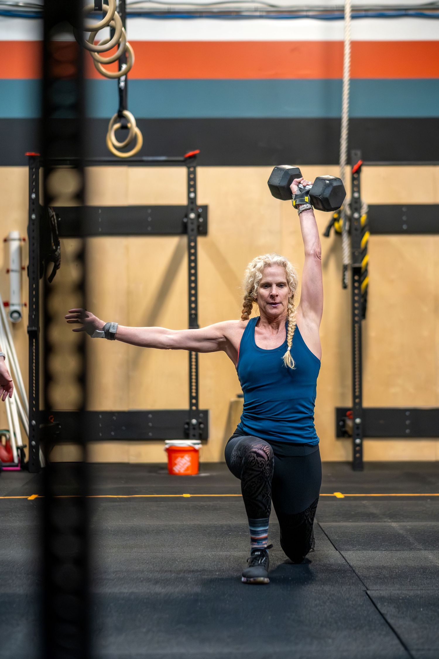 Fit blonde woman performs a kneeling overhead dumbbell press in a CrossFit gym setting.