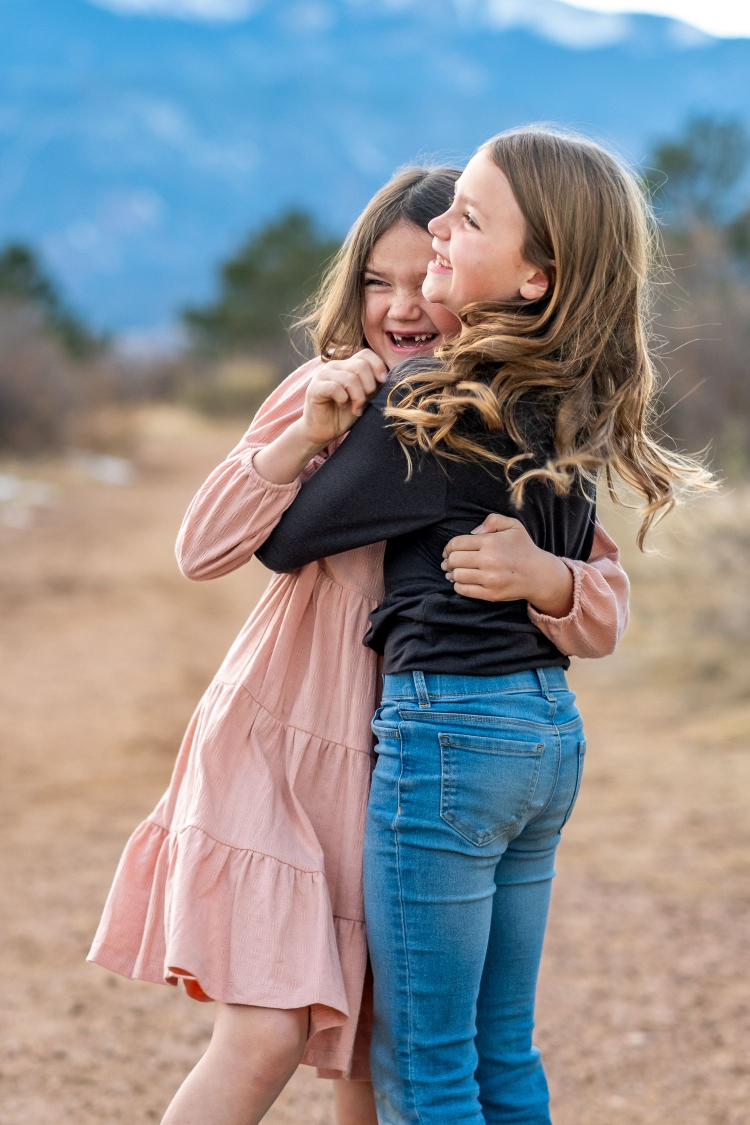 Two young girls laughing and hugging each other outdoors with mountains visible in the background.
