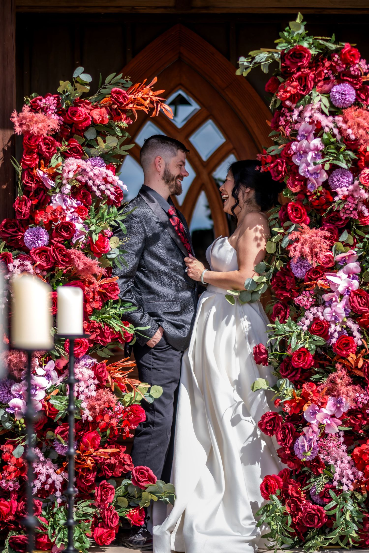 Joyful couple stands beneath stunning red and pink floral arch at wooden chapel wedding venue.