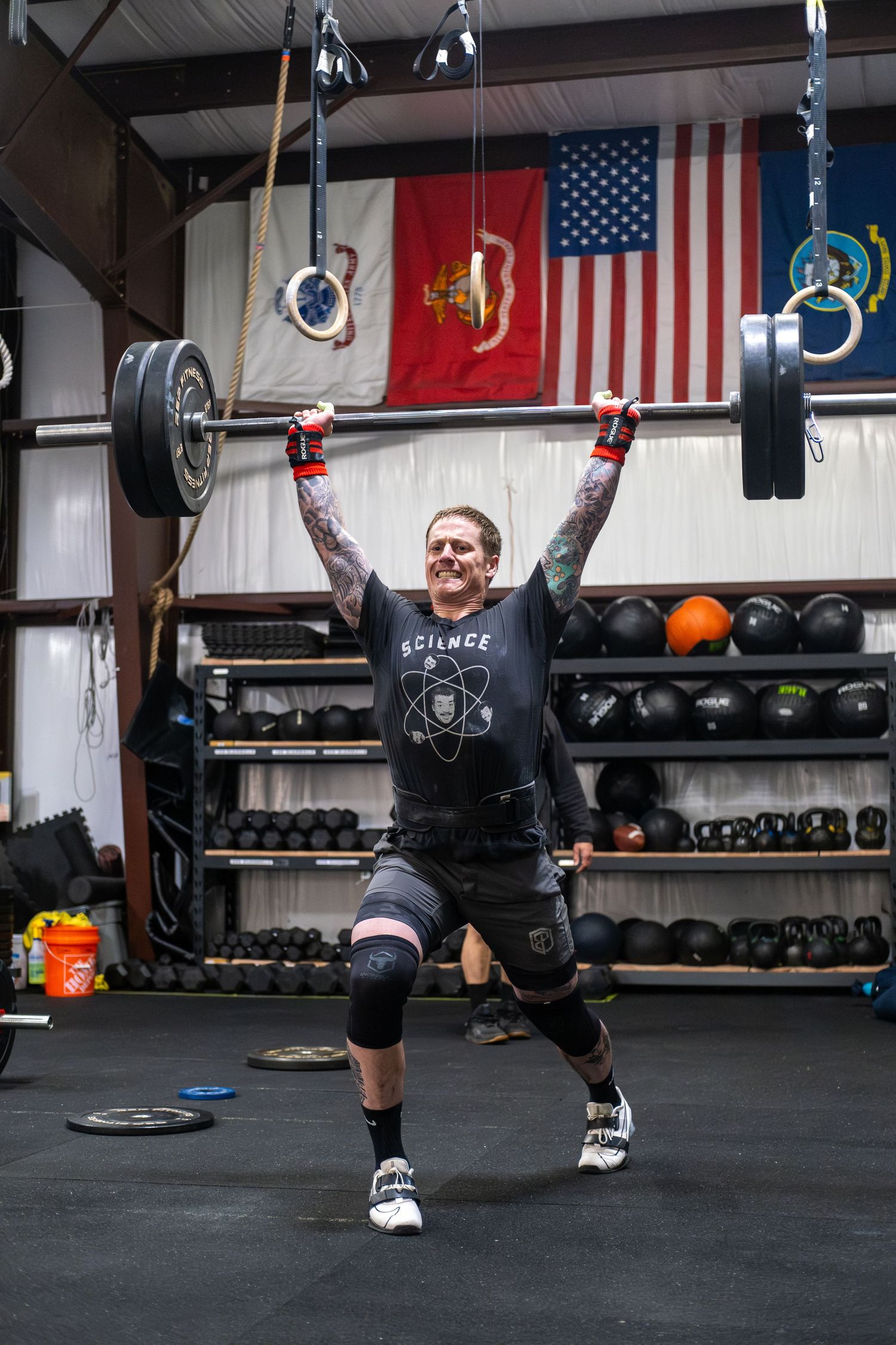 Tattooed man in black shirt completes an overhead barbell lift in a gym with American flags on wall.