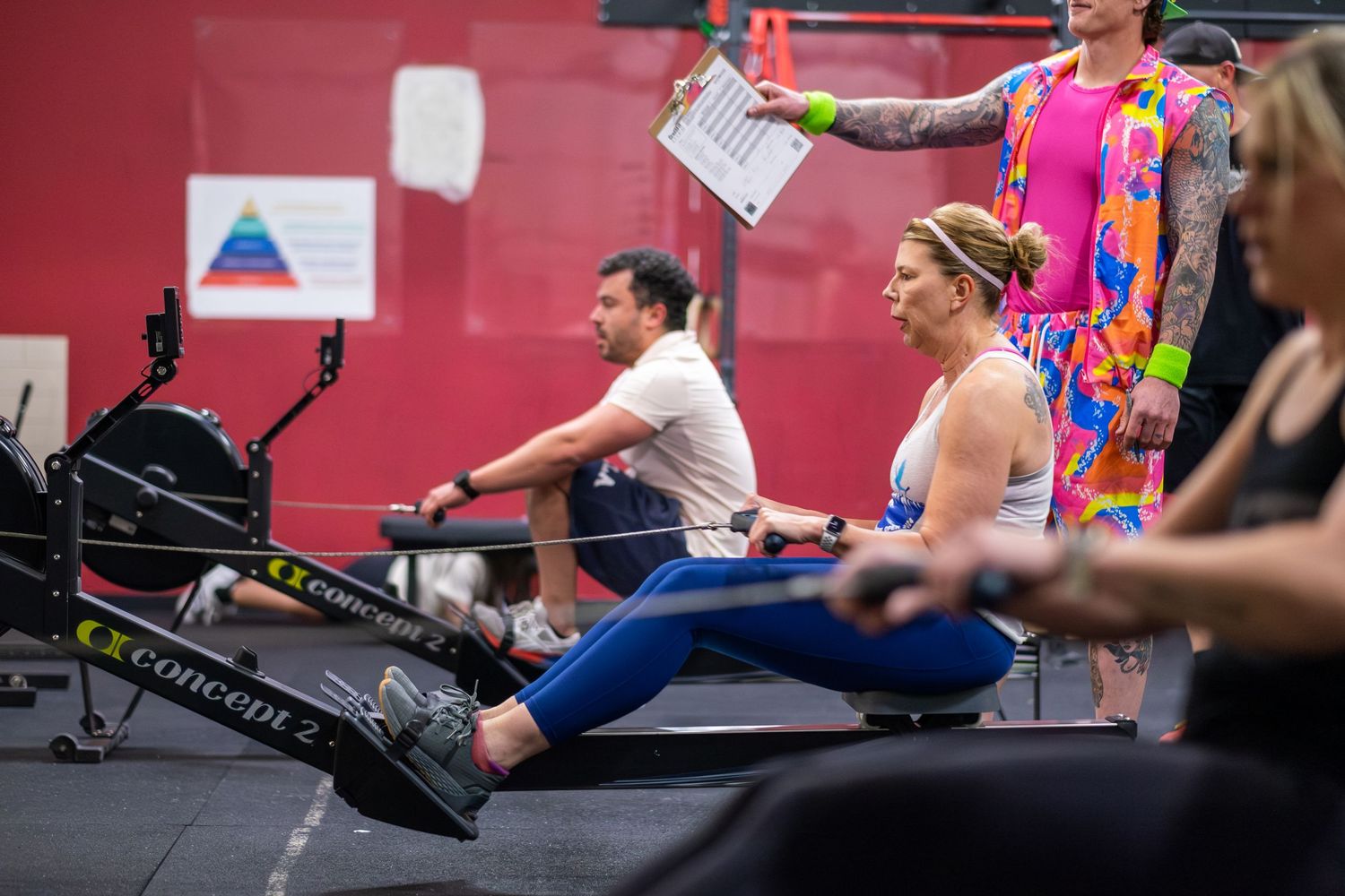 Athletes row on Concept2 rowing machines during a CrossFit competition while a judge observes.