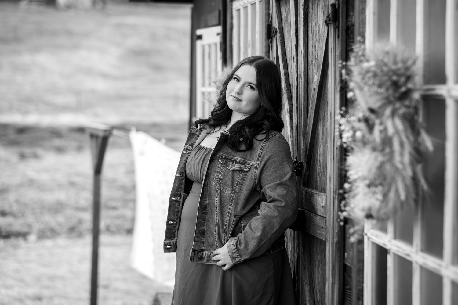 Black and white portrait of a woman in a denim jacket leaning against a rustic wooden barn door.