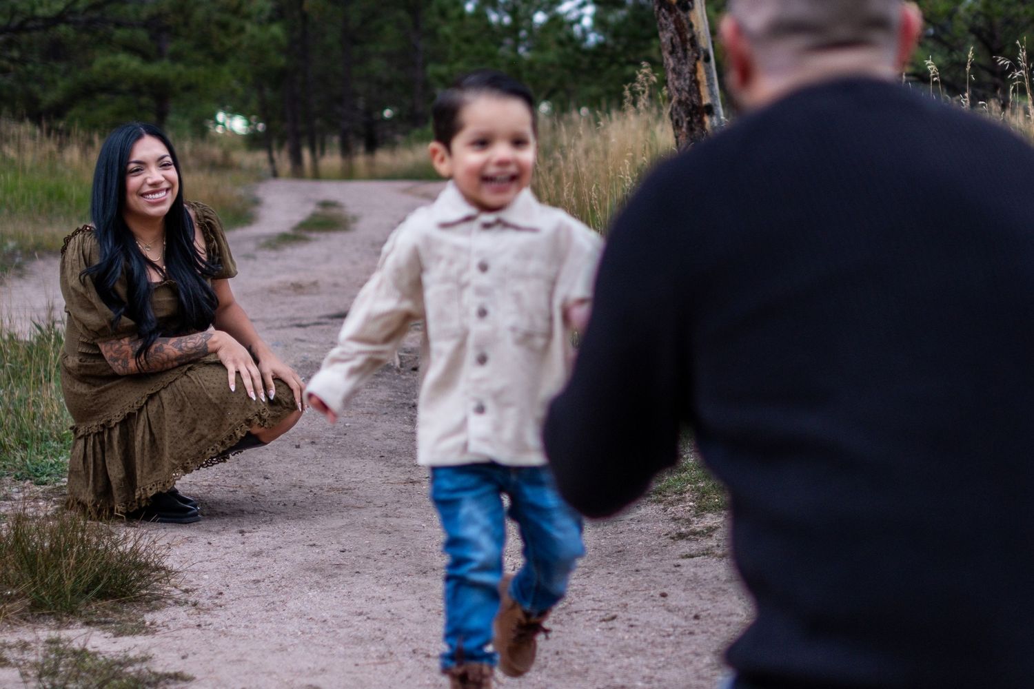 Young boy running toward father on dirt path while mother smiles in pine forest.