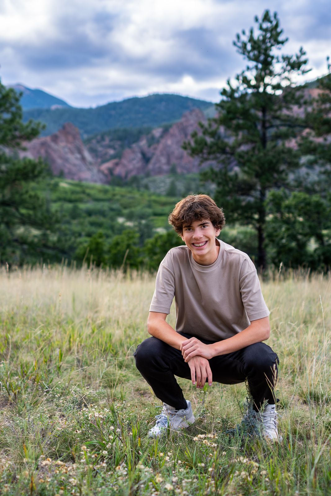 Smiling teenage boy crouching in a Colorado mountain meadow with pine trees and rocky peaks behind him.