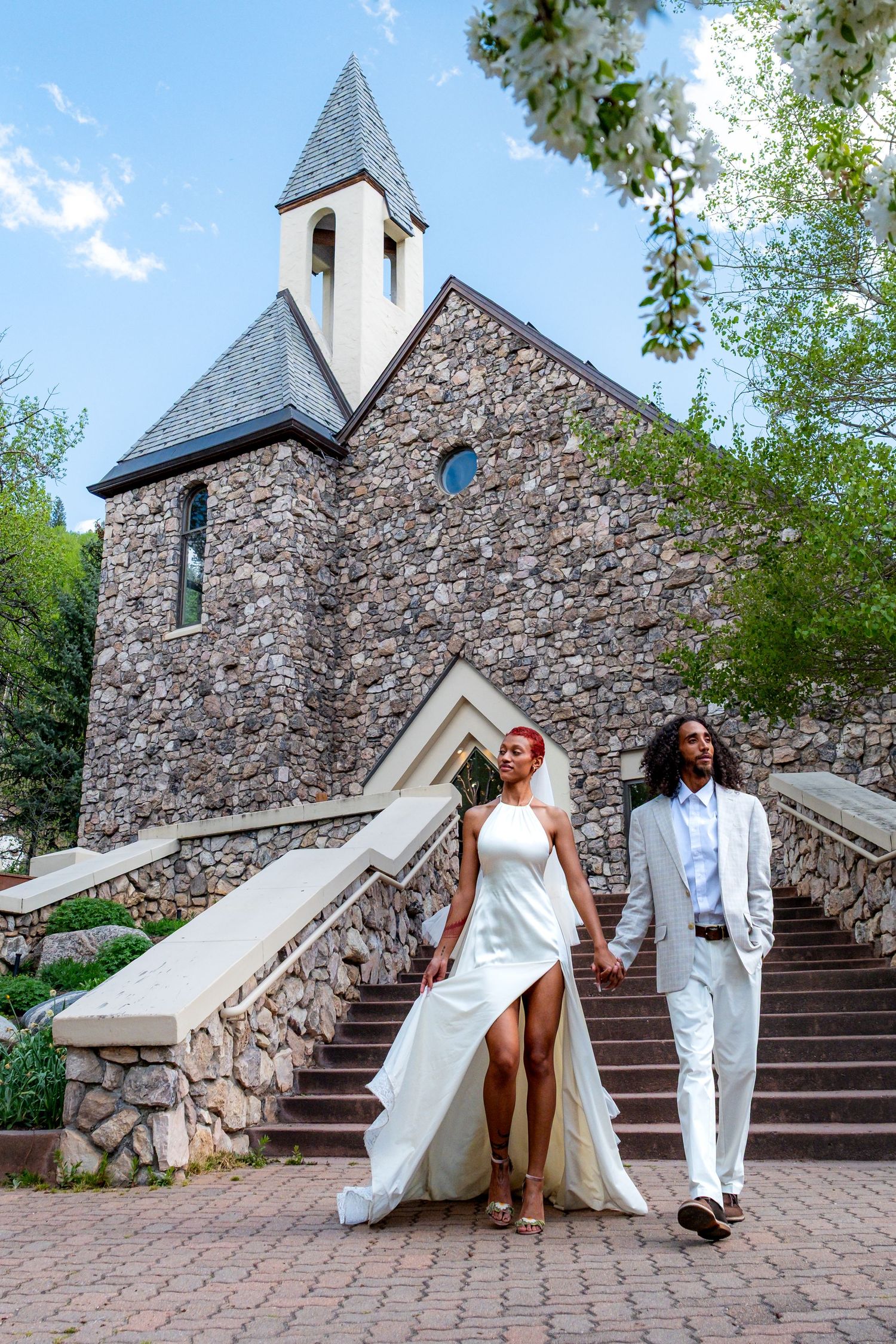 Newlywed couple walking hand in hand down steps of stone chapel surrounded by blooming trees.
