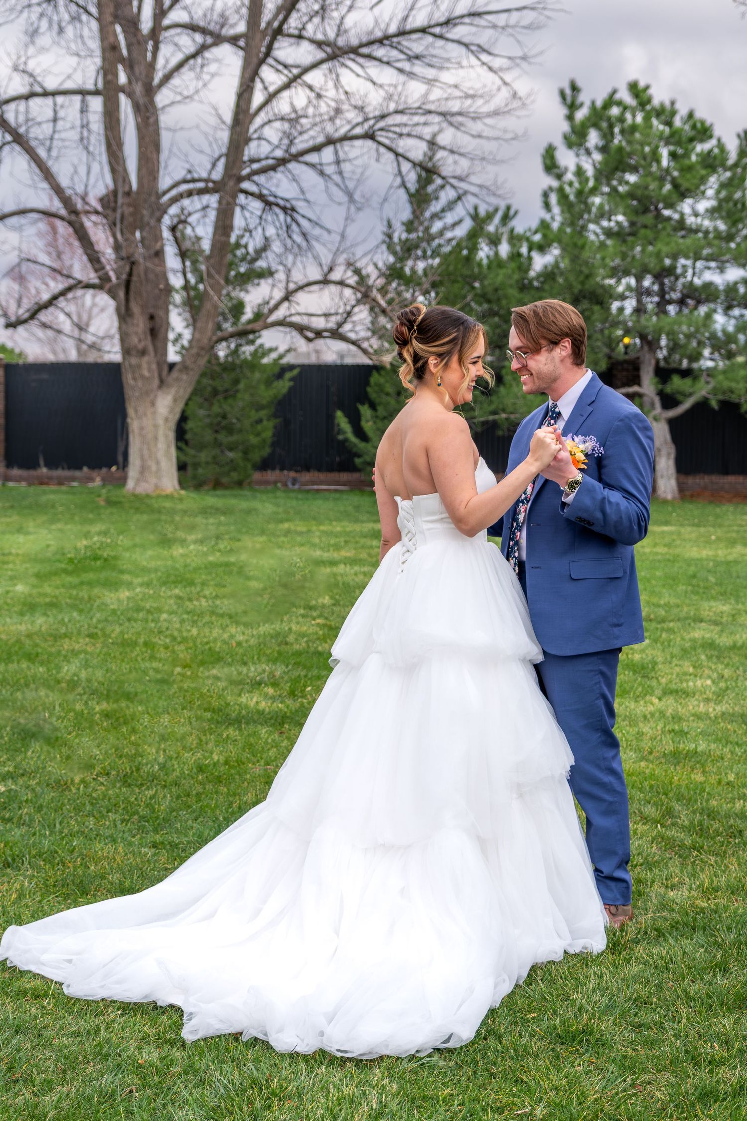 Bride and groom share a tender moment on green lawn, groom in blue suit holding colorful bouquet.