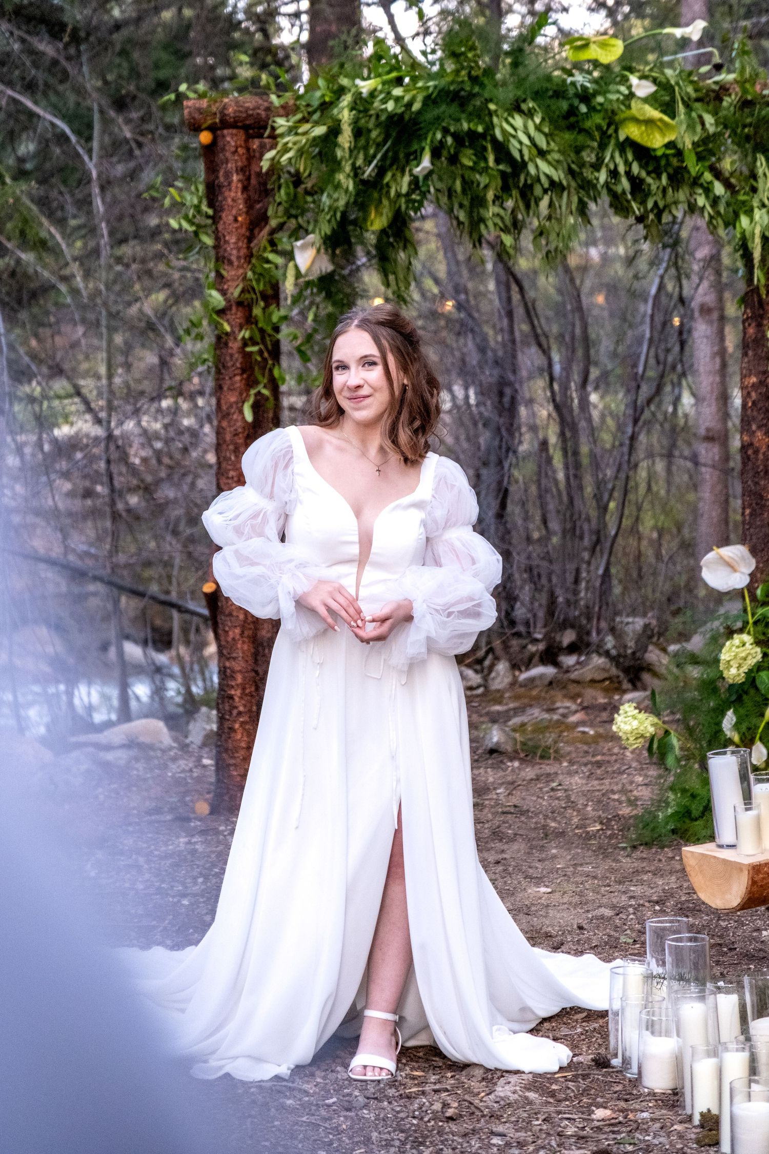 Elegant bride in white puff-sleeve gown with slit standing under rustic floral arch surrounded by candles in forest.