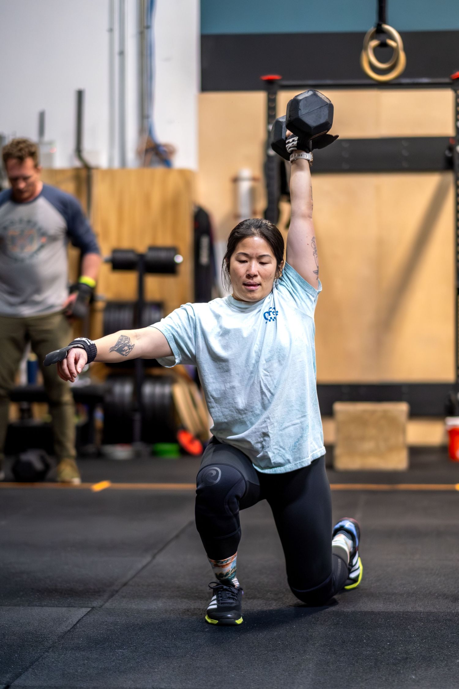 Asian woman performs a kneeling single-arm dumbbell overhead hold during a CrossFit workout.