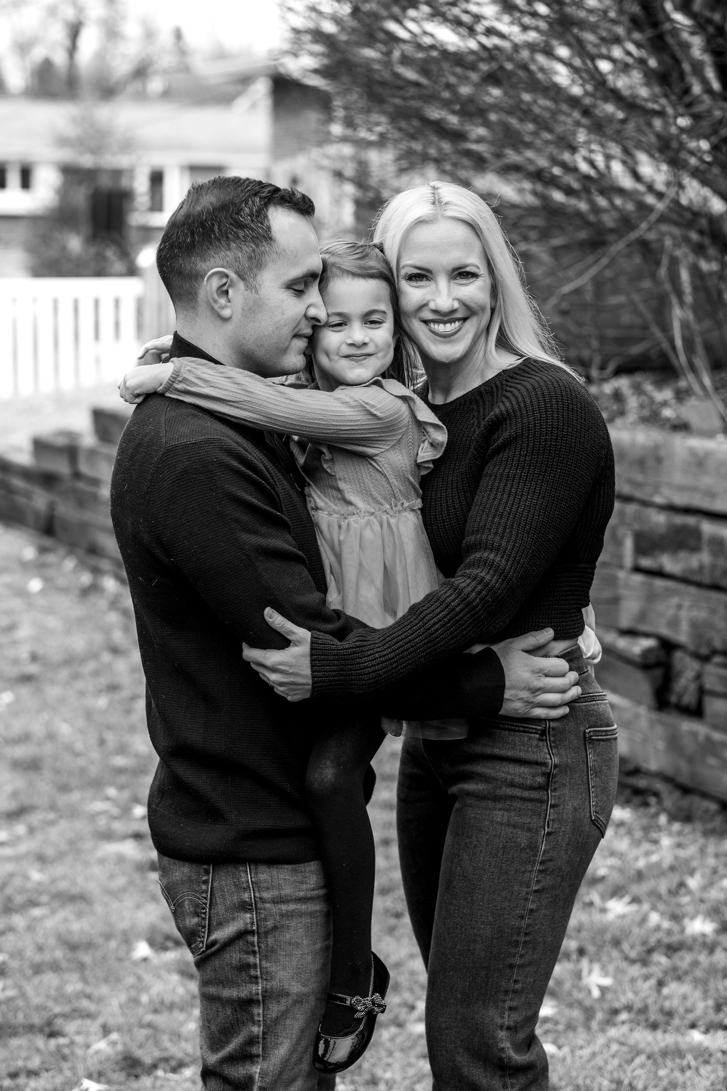 Black and white portrait of loving family, parents embracing young daughter in outdoor garden setting.