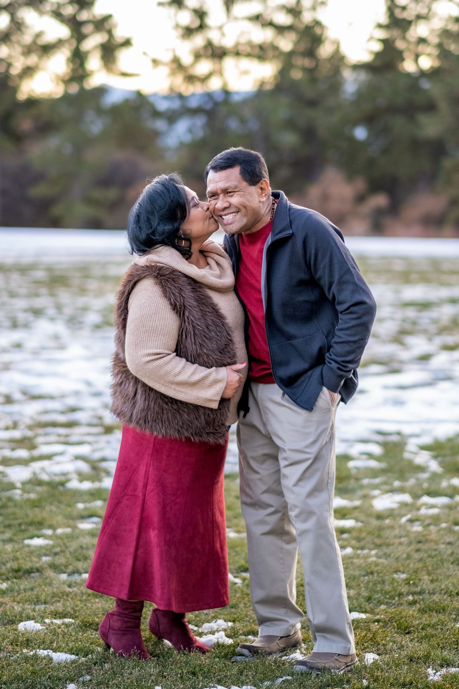 Couple sharing a romantic kiss outdoors in a snowy winter park setting, woman in red skirt.