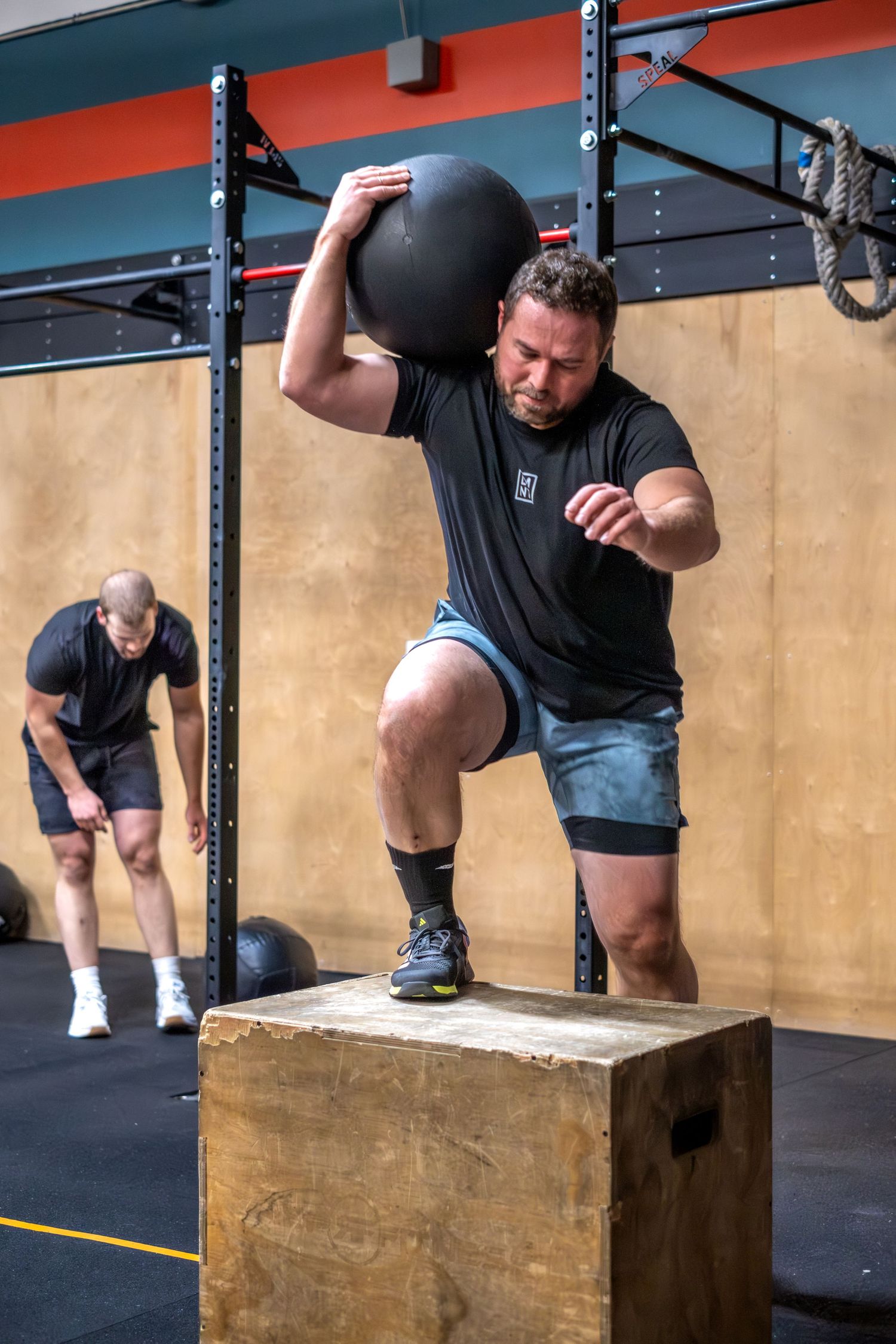Man performing a box jump with a medicine ball overhead in a CrossFit gym workout.