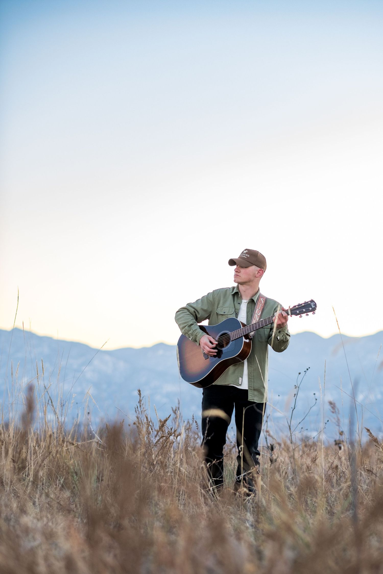 Young man playing acoustic guitar in golden field with mountain range visible in background at sunset.
