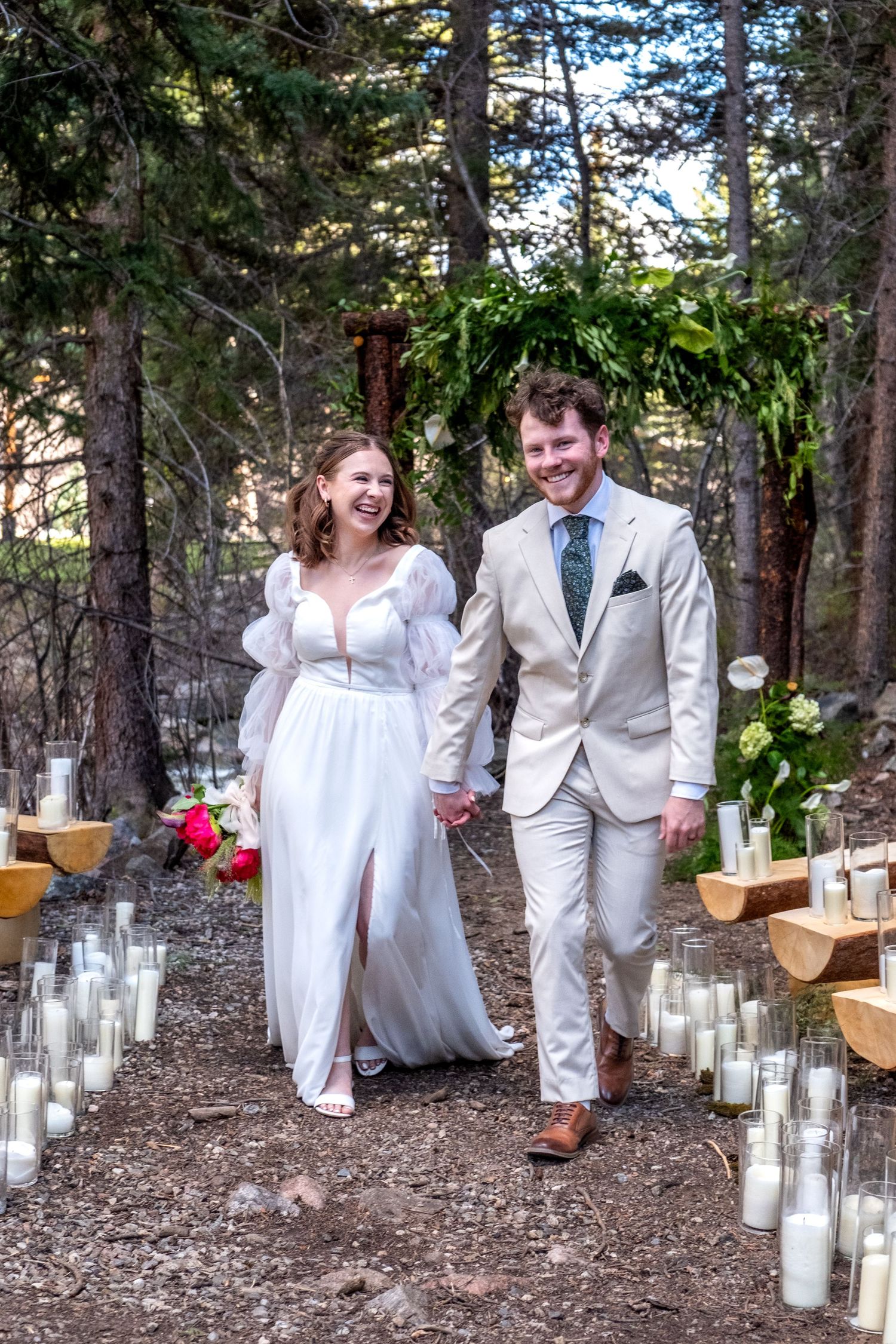Smiling newlyweds walking hand-in-hand down candle-lined aisle in forest, bride in puff-sleeve gown, groom in tan suit.