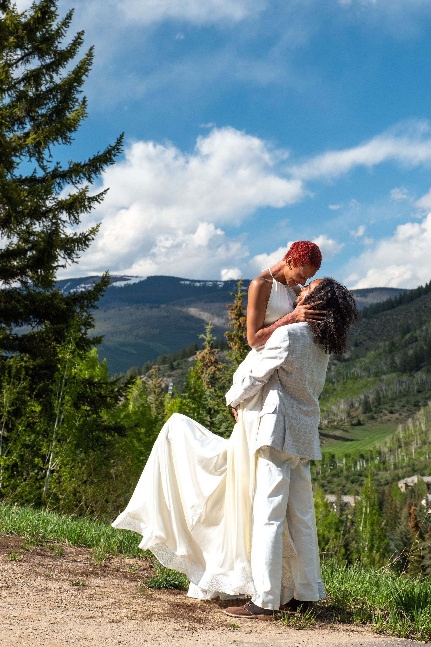 Groom lifting bride in white wedding dress against scenic Colorado mountain landscape on sunny day.