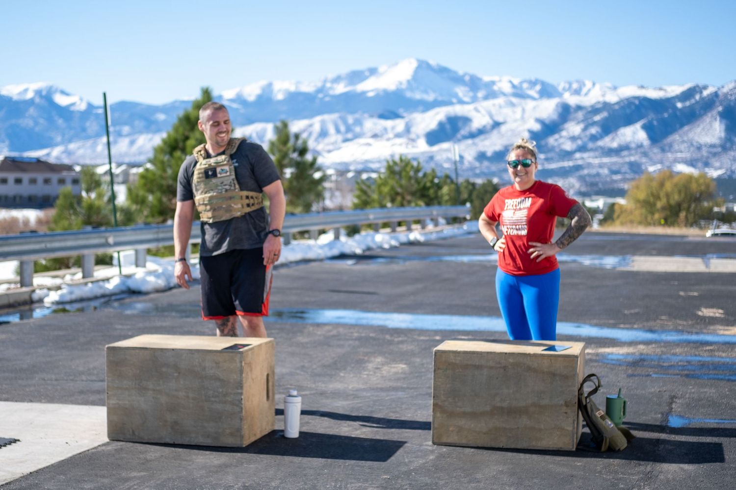 Two athletes stand on wooden plyo boxes outdoors with snow-capped mountains in the background.
