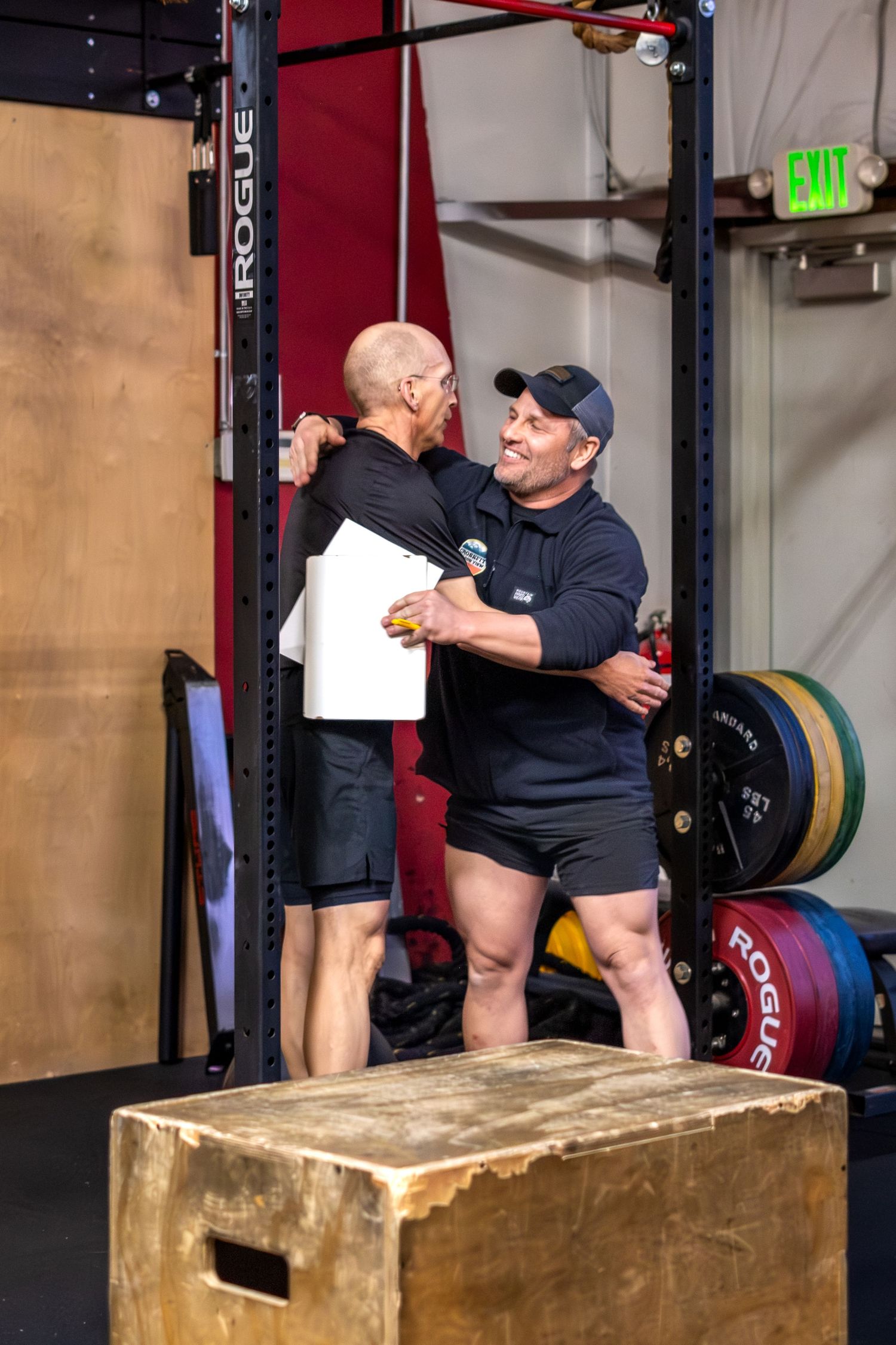 Two men embracing and congratulating each other in a CrossFit gym near a barbell rack.