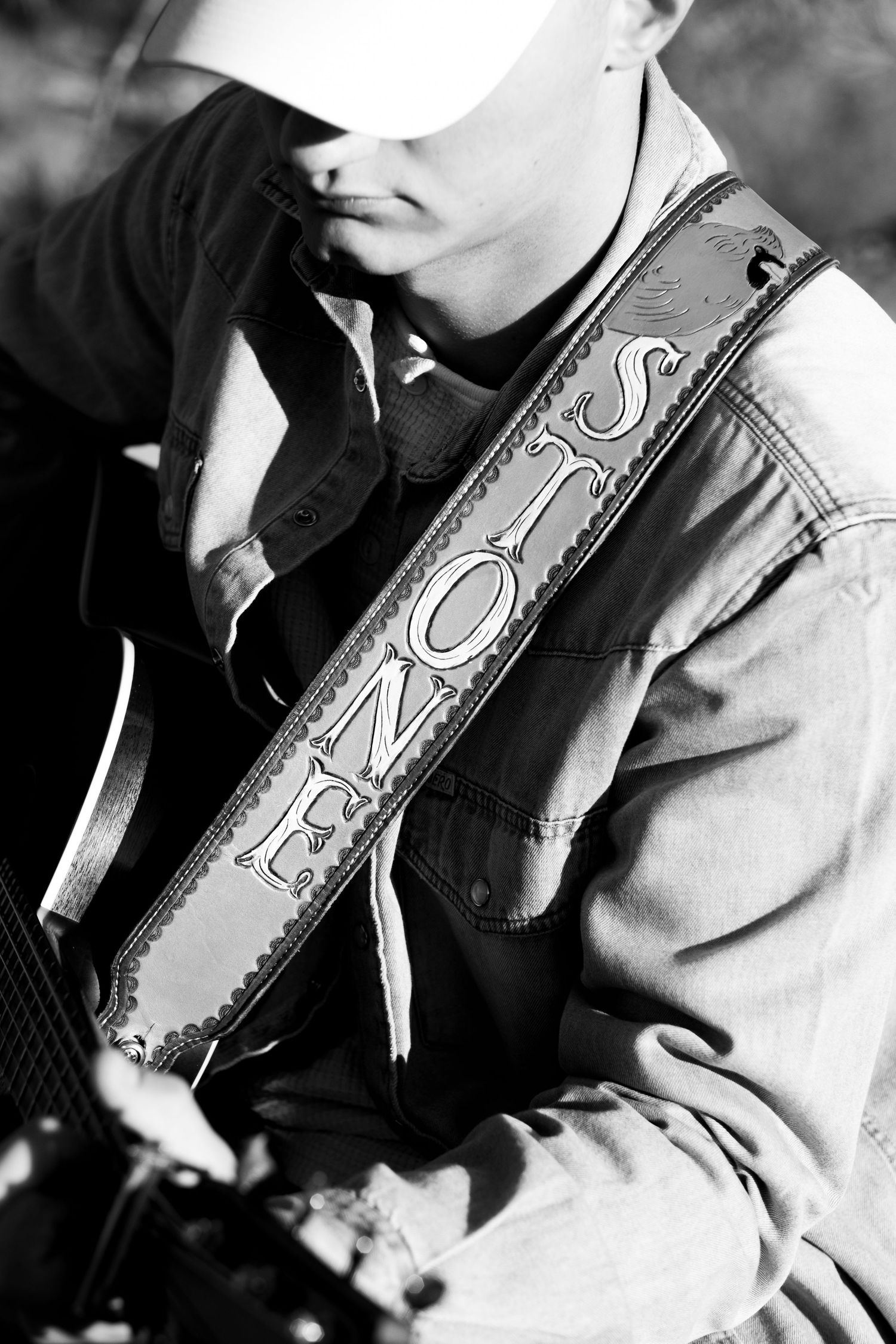 Black and white close-up of guitarist with decorative leather guitar strap reading Stone outdoors.