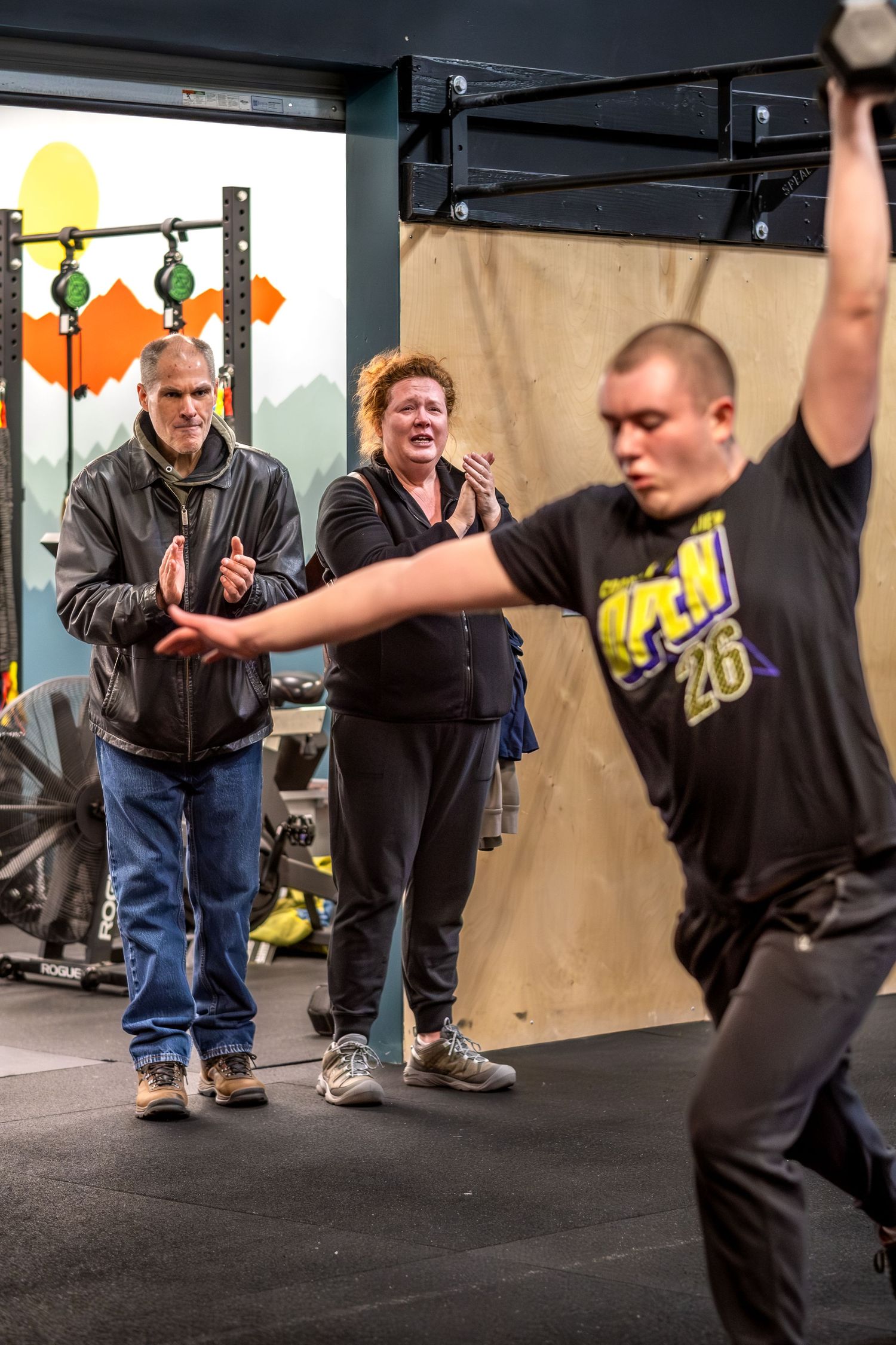 Two spectators cheer as a young man performs an overhead dumbbell lift at a CrossFit event.