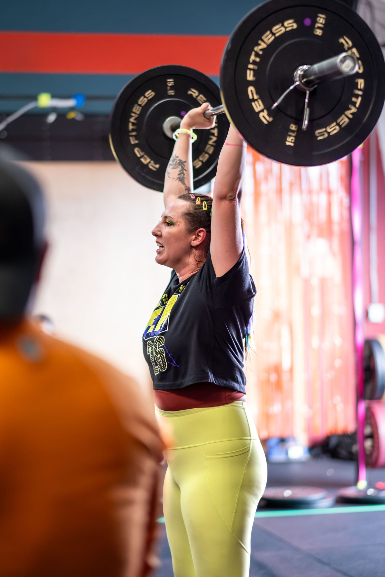 Woman in yellow leggings lifting heavy barbell overhead at a CrossFit competition.