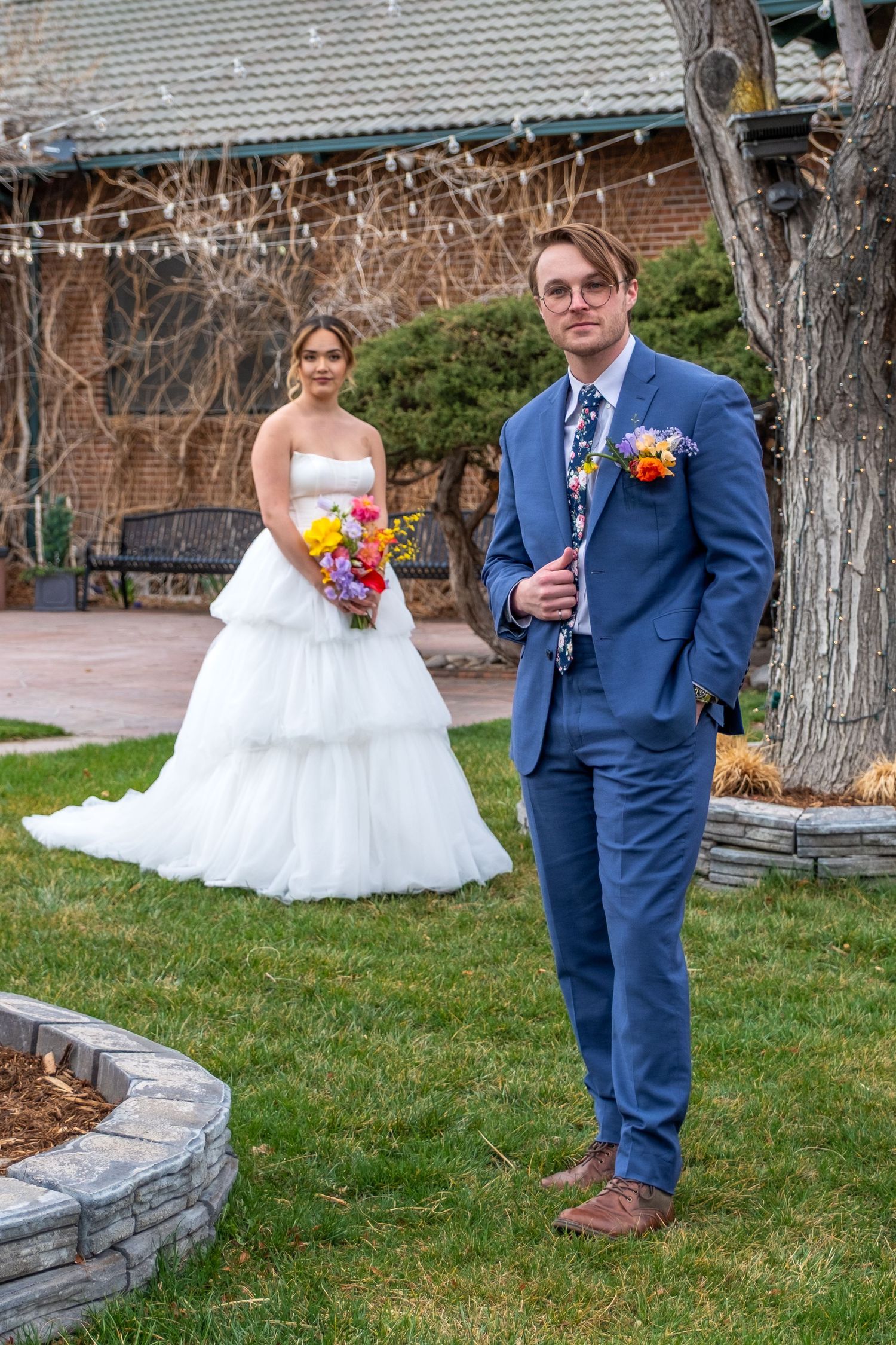 Bride in tiered white gown and groom in blue suit pose outdoors at garden wedding venue.